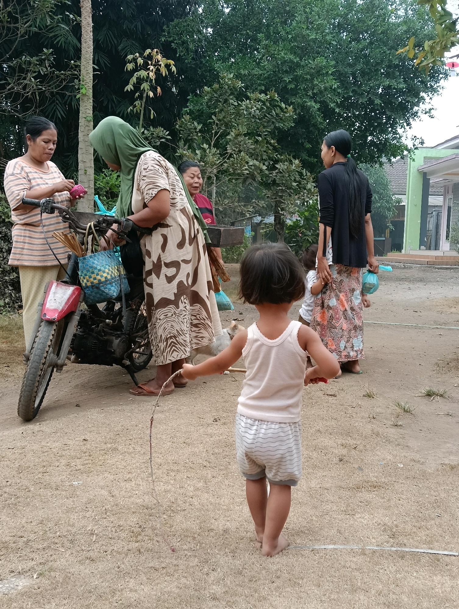 In Kasiyan Timur, Indonesia, a group of people are gathered near a motorcycle. Two adult women are standing to the left of the motorcycle, one wearing a striped t-shirt and the other a patterned dress and green headscarf. They appear to be interacting with items on the motorcycle, possibly loading or unloading. A third adult woman stands further back, partially obscured by trees. In the foreground, a young child with dark hair is facing away from the camera, holding a jump rope. Another small child is visible near the taller woman in the black shirt and patterned skirt. The environment is outdoors, with dry, sandy ground and lush green trees forming a backdrop. The buildings in the background suggest a residential area.