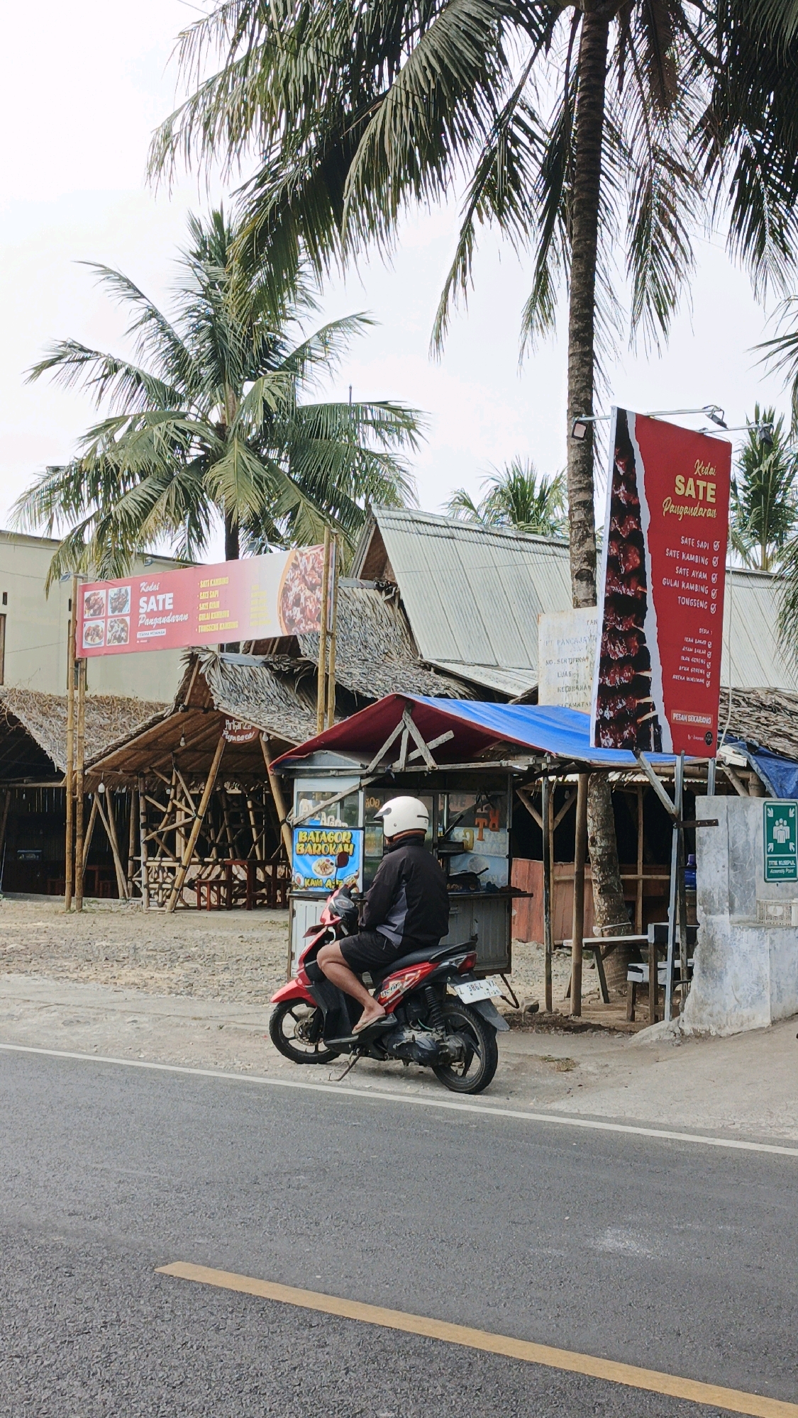 The image depicts a street scene in Desa Wonoharjo, Indonesia, with a focus on a roadside food stall and a person on a motorcycle. The environment is characterized by palm trees and rustic, open-air structures that appear to be food stalls or small eateries with thatched roofs and bamboo supports. A prominent red banner with white text hangs from a pole, advertising "Kedai Sate Panggarauran" and listing various sate dishes like "Sate Sapi," "Sate Kambing," and "Sate Ayam." Another banner, to the left, also advertises "Sate Panggarrauran" with images of food.
In the foreground, a person wearing a black jacket, black shorts, and a white helmet is seated on a red motorcycle. The motorcycle is parked on the side of a paved road, with a yellow line marking the lane. The person's right foot is resting on the ground with a sandal. Behind the motorcycle and the person is a food stall with a blue tarp roof and various items displayed, including a sign for "BATAGOR BAROKAH." The stall has a grey metallic structure. In the background, more of these thatched-roof structures are visible, along with a building with a metal roof. The sky is overcast.