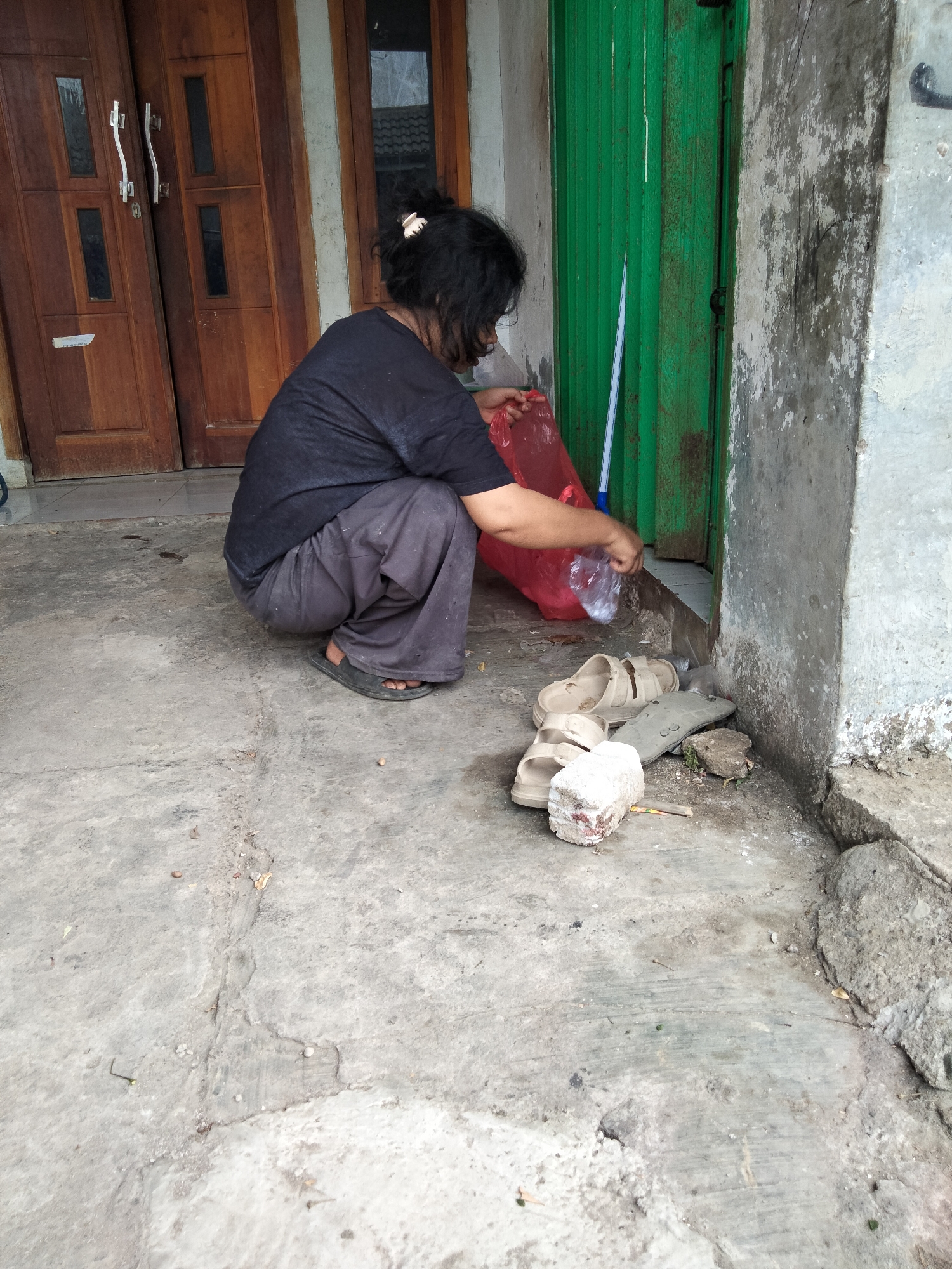 A person is squatting on a concrete surface in front of a wooden door and a green corrugated metal door. The person is wearing a dark t-shirt and dark pants, and is holding a red plastic bag and a clear plastic bottle. There are several pairs of sandals and a piece of white brick on the ground near the person. The scene appears to be outdoors, possibly in a residential or commercial area. The location is identified as Cariu, Indonesia.