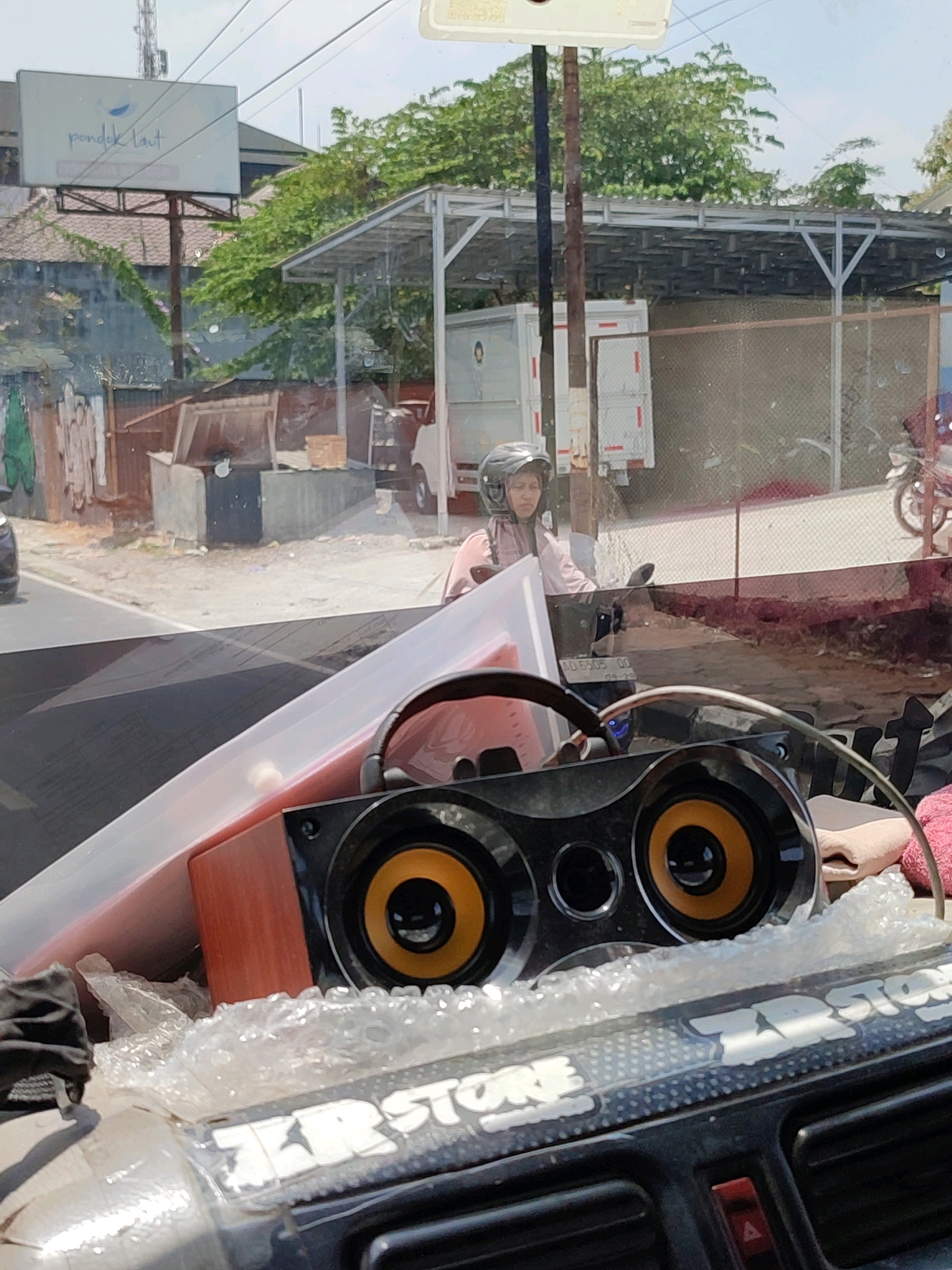 The image shows a scene viewed through a vehicle's windshield, possibly in Magelang, Indonesia. In the foreground, a black speaker with orange accents and a wooden casing is visible, partially wrapped in bubble wrap. The dashboard of the vehicle is also present, with a red button and air vents.
Beyond the vehicle, a woman wearing a helmet and a light pink jacket is riding a motorcycle. She is looking forward. Behind her, a white truck with a closed cargo area is parked. In the background, a tall cell tower stands against a bright sky. A billboard with blue text and a wave graphic is also visible, mounted on a metal structure. Trees with green foliage are abundant, and there's a fenced area with a metal roof structure in the middle ground. The overall environment appears to be a street or an area with commercial or industrial buildings.