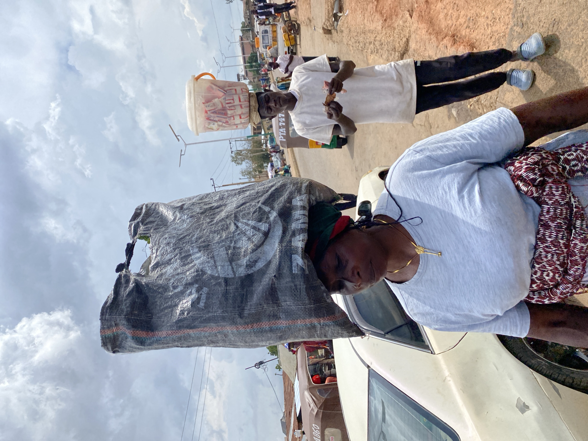 The image depicts a street scene in Bukuru, Nigeria, under a cloudy sky. Two individuals are the primary subjects. In the foreground, a person is partially visible, wearing a light grey t-shirt and a patterned cloth around their waist. They are leaning against a cream-colored car, with a large, dark-colored woven sack resting on their head and shoulders, obscuring much of their body. The sack appears to be made of rough material and is tightly packed.
Behind this person, another individual is seen walking, carrying a clear plastic bucket filled with packaged goods on their head. This person wears a white t-shirt and dark pants, and is holding a small, wrapped item in their right hand, possibly food. They are positioned near a three-wheeled vehicle.
The background reveals a busy street with other vehicles and people, suggesting a marketplace or a bustling thoroughfare. The overall impression is one of daily life and commerce. The weather appears overcast, indicating it might be a humid or potentially rainy day. No specific time of day is discernible, but the lighting suggests it's daytime. The setting is clearly an outdoor urban environment in Nigeria. The emotions of the individuals are not clearly discernible, though the person in the foreground seems to be looking down or to the side, while the person in the background is looking towards the camera. Visible text includes "PTA: 747" on the side of the three-wheeled vehicle.
