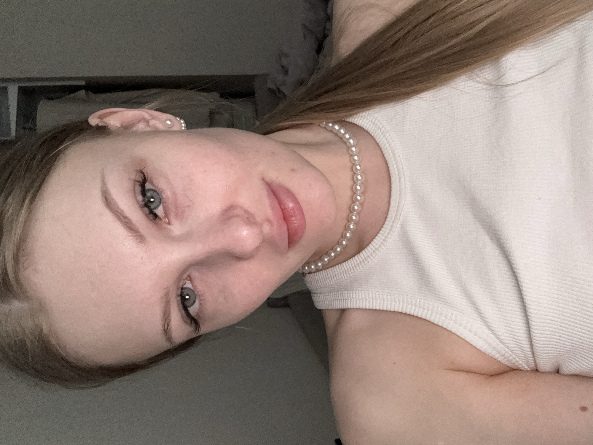 A young woman with blonde hair and fair skin is shown in a close-up selfie. She is wearing a white ribbed tank top and a pearl necklace. She has light-colored eyes and appears to be wearing minimal makeup, with a focus on her eyelashes. She also has a pearl earring. The background is indistinct and dark, with some furniture visible. The photo does not provide information about the location being the city of Perm, Russia.