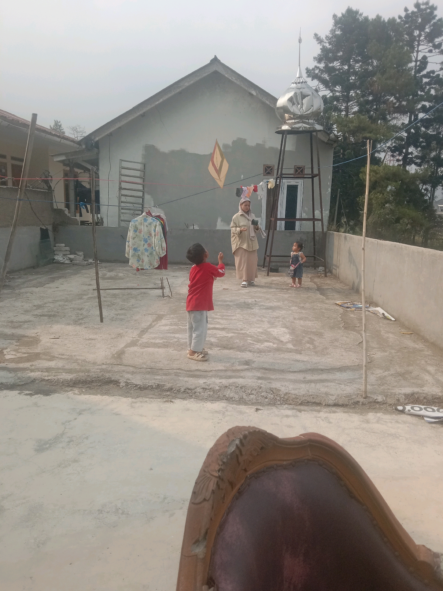 In Sukanagalih, Indonesia, an outdoor rooftop scene unfolds with three people and various objects against a backdrop of a partially constructed building and lush trees. The sky is overcast, casting a diffused light over the environment.
In the foreground, a child wearing a red long-sleeved shirt and light-colored pants is positioned in the center-left, facing away from the camera and looking upwards. To their right, a woman in a light-colored hijab and long skirt stands, holding an object in her right hand. A smaller child, dressed in a dark denim overall with a floral print top and pink sandals, stands to the woman's right.
The rooftop itself is made of concrete with visible expansion joints and patches of repair. A clothesline extends across the area, holding several garments, including a floral-patterned shirt and a red garment. Two wooden poles support the clothesline. A metal structure, possibly a water tank, is visible to the left of the woman, topped with a decorative silver dome that resembles a mosque's finial. A kite with diamond-shaped patterns in yellow and red is attached to a string and hangs on the side of the building.
The building behind the people is a two-story structure with gray walls and a gray tiled roof. Windows and doors are visible. To the right of the building, a dense cluster of tall green pine trees rises against the sky. In the bottom right corner of the image, the edge of a dark red upholstered chair with an ornate wooden frame is visible, suggesting the vantage point of the photographer. Debris, including what appears to be construction materials and a flattened mat or rug, is scattered on the rooftop. The overall impression is a domestic scene on a rooftop, possibly in a residential area, during the day.