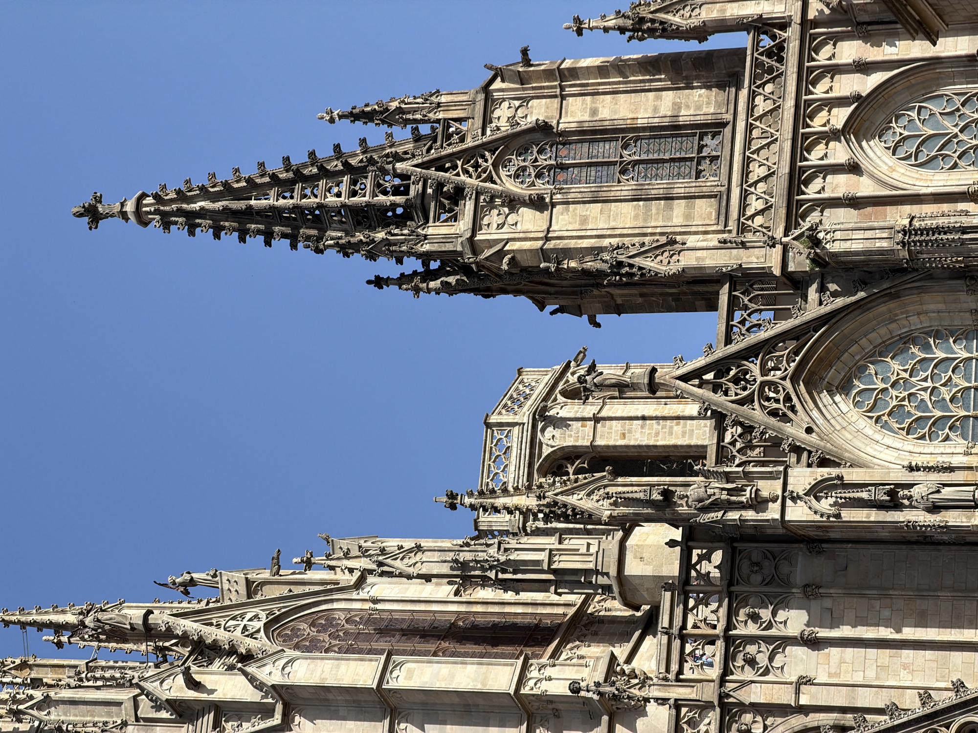 This is a vertical, eye-level shot of the Barcelona Cathedral in Barcelona, Spain. The image focuses on the intricate Gothic architecture of the cathedral's facade, with a clear blue sky as the background. Sunlight casts shadows, highlighting the stone carvings, spires, and gargoyles. Two prominent spires dominate the right side of the frame, one taller than the other. Numerous decorative elements, including pointed arches, tracery, and statues, adorn the stonework. The photograph captures the grandeur and detail of the cathedral's exterior.