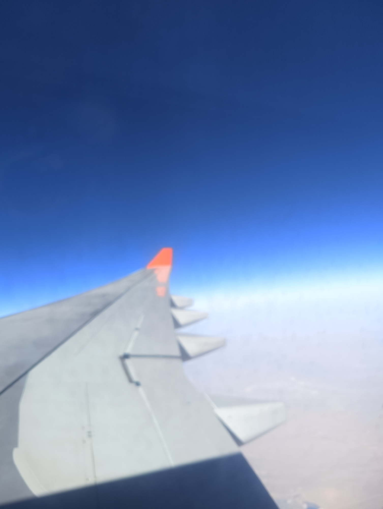 An airplane wing is visible against a deep blue sky. The wing is a light gray color with some white elements and an orange tip. Below the wing, there are faint hints of clouds and possibly land. The image is out of focus, giving it a blurred appearance. There are no people or specific landmarks identifiable within the frame. The location in Nakhabino, Russia, cannot be determined from the visual information provided.