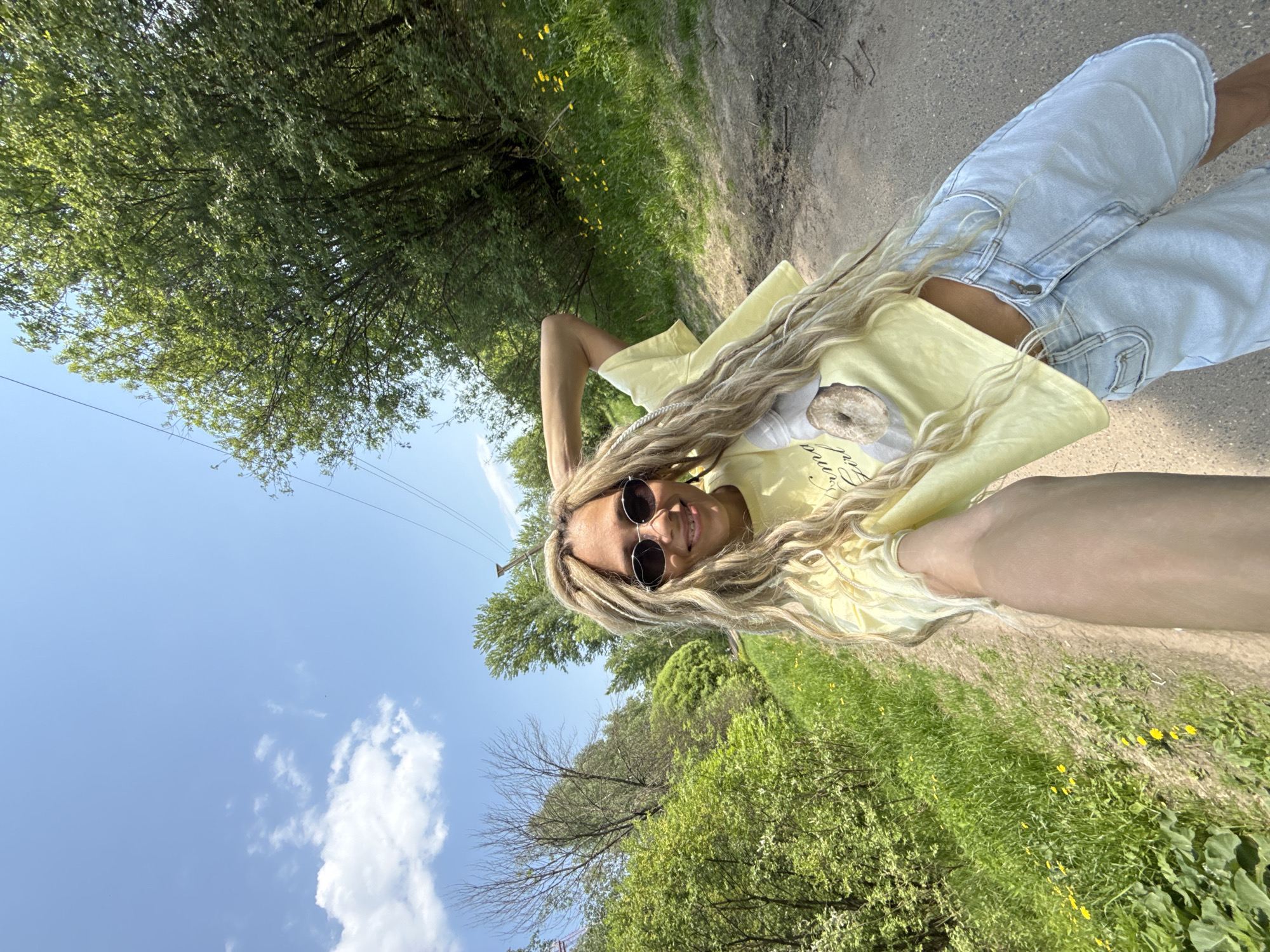 A photograph captures a woman, presumably posing for a selfie, on what appears to be a dirt path through a wooded or grassy area. The woman is wearing light-colored denim shorts and a yellow t-shirt with a graphic of a donut. She has long, light-colored hair, likely dreadlocks, and is wearing round, dark sunglasses. She is smiling, and her head is slightly tilted.
The environment consists of a dirt path flanked by grass and trees. The sky is visible in the upper-left corner with some clouds. It's a bright, sunny day. The image is angled, giving the impression of someone lying on the ground taking a photo from a low perspective. There are power lines overhead on the left.
I am unable to verify whether the photograph was taken in Rusanovo, Russia.
