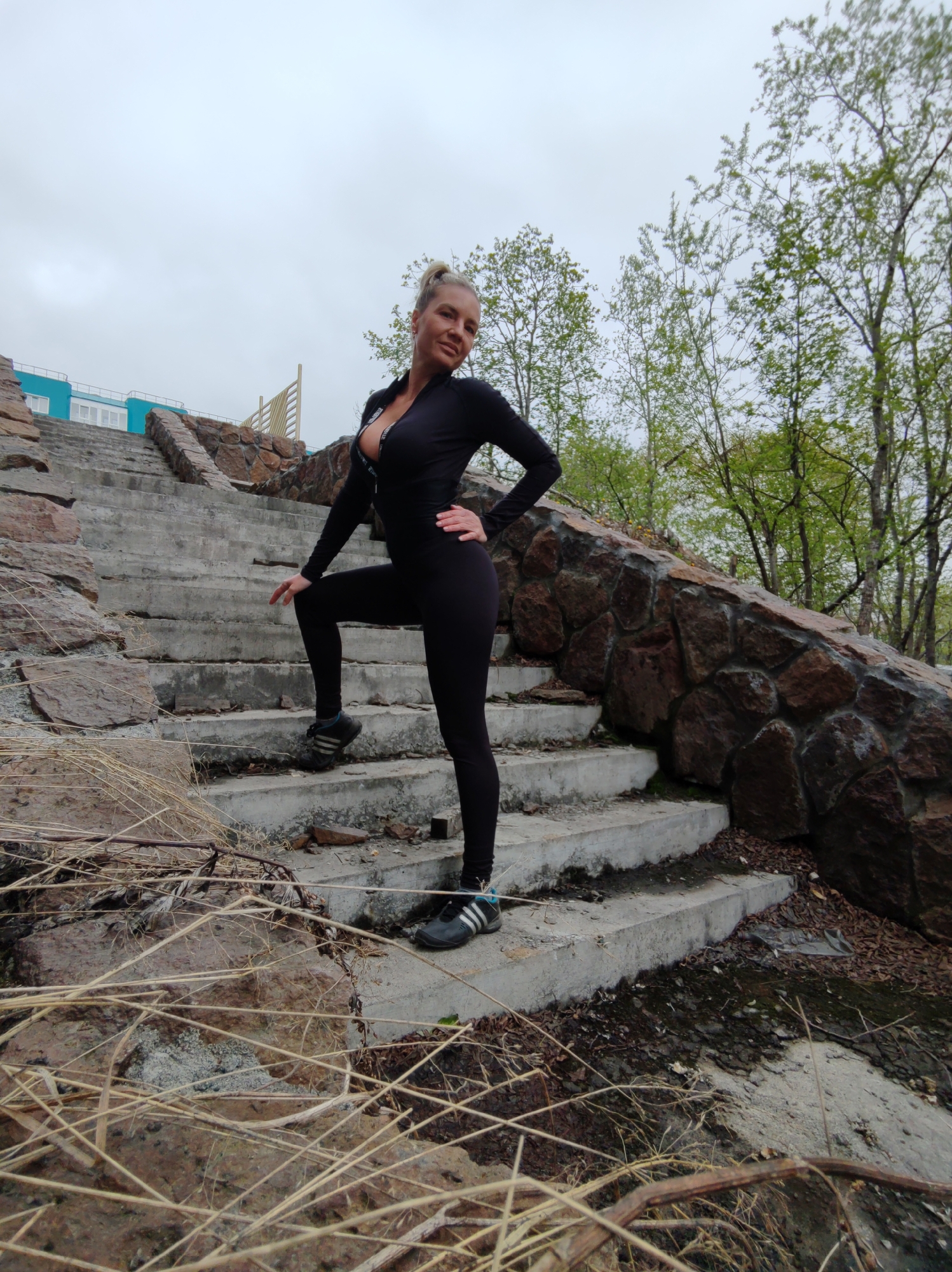 A woman in a black athletic jumpsuit stands on a concrete staircase in Petropavlovsk-Kamchatsky, Russia. The staircase is part of an outdoor structure, possibly a park or public space, with a stone wall on the right side and a light blue building in the background to the left. Trees with green leaves are visible behind the woman and the stone wall. Dry grass and fallen leaves are scattered on the ground and on the stairs, indicating an outdoor, possibly somewhat unkempt, environment. The sky is overcast. The woman is posing with one hand on her hip and the other resting on the stair.