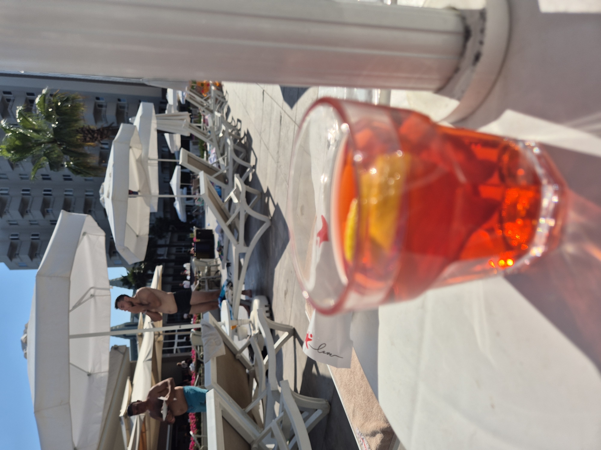 The image depicts a poolside scene on a sunny day. The perspective is from a person holding a red drink in a clear glass, which is prominently featured in the foreground. The drink contains ice, citrus slices, and appears to be a cocktail. Behind the glass, a white cloth with the word "love" written in script is visible, possibly a napkin or a piece of clothing.
In the middle ground, several people are visible, some in swimwear. One man in black swim trunks is standing and looking to his left. Another person in blue swim trunks is sitting. There are numerous white lounge chairs and umbrellas arranged on a paved area, suggesting a resort or hotel setting. A palm tree can be seen on the left side of the image, further indicating a warm climate.
In the background, a multi-story building with many windows is visible. The overall atmosphere appears relaxed and leisurely, consistent with a vacation or a day spent relaxing outdoors. The bright sunlight casts distinct shadows, indicating it is likely daytime. The image is oriented vertically, with the top of the frame on the left.