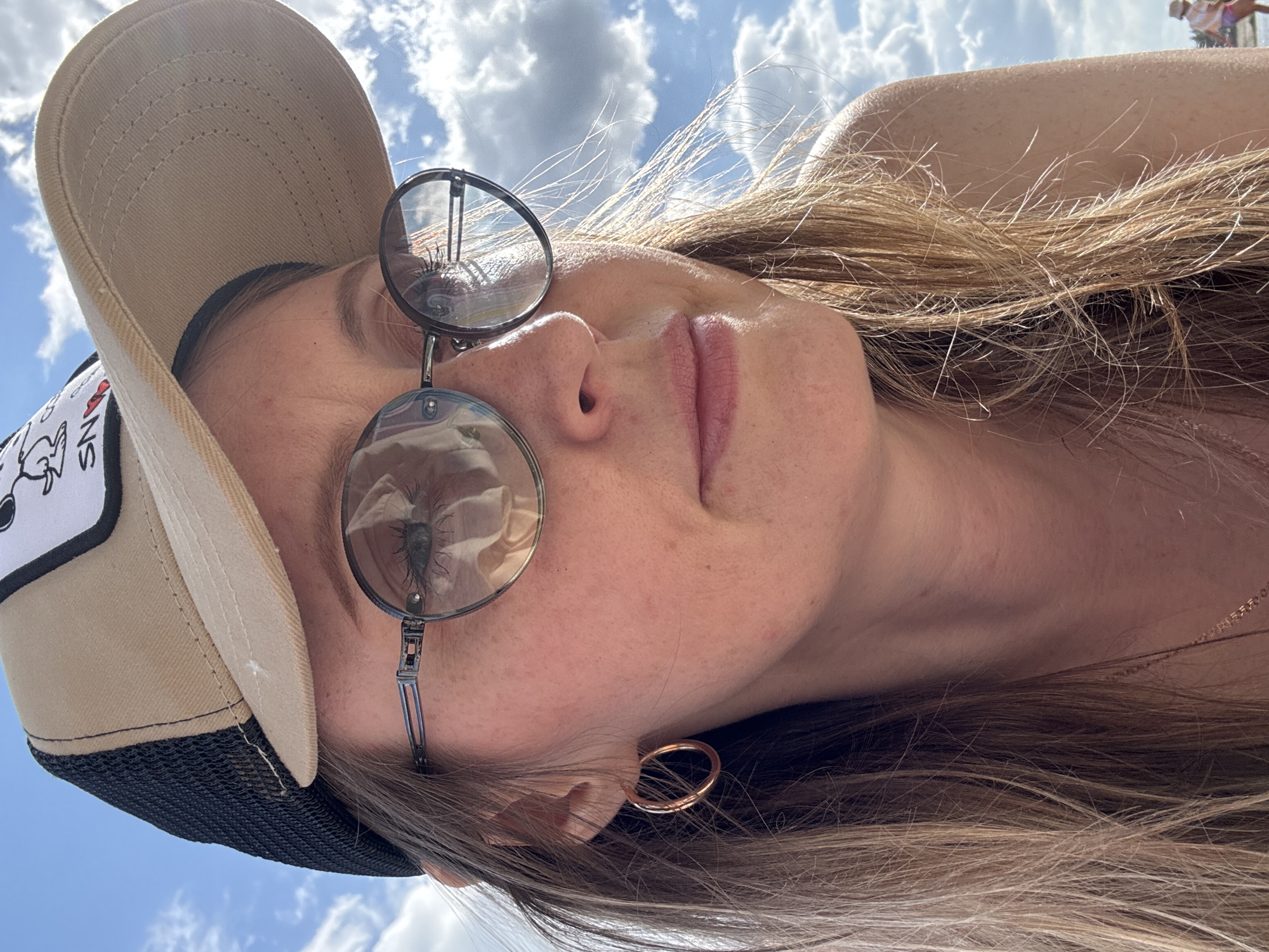 A woman is seen from a low angle, tilting her head back towards the sky. She is wearing a beige baseball cap with a black mesh backing and a small patch on the front. Her hair is long, blonde, and straight. She has round, metal-framed glasses on, and a gold hoop earring in her left ear. The reflection in her glasses shows details of the sky and possibly a bug. The background shows a cloudy sky. The image was taken outside in the city of Chaikovsky, Russia.