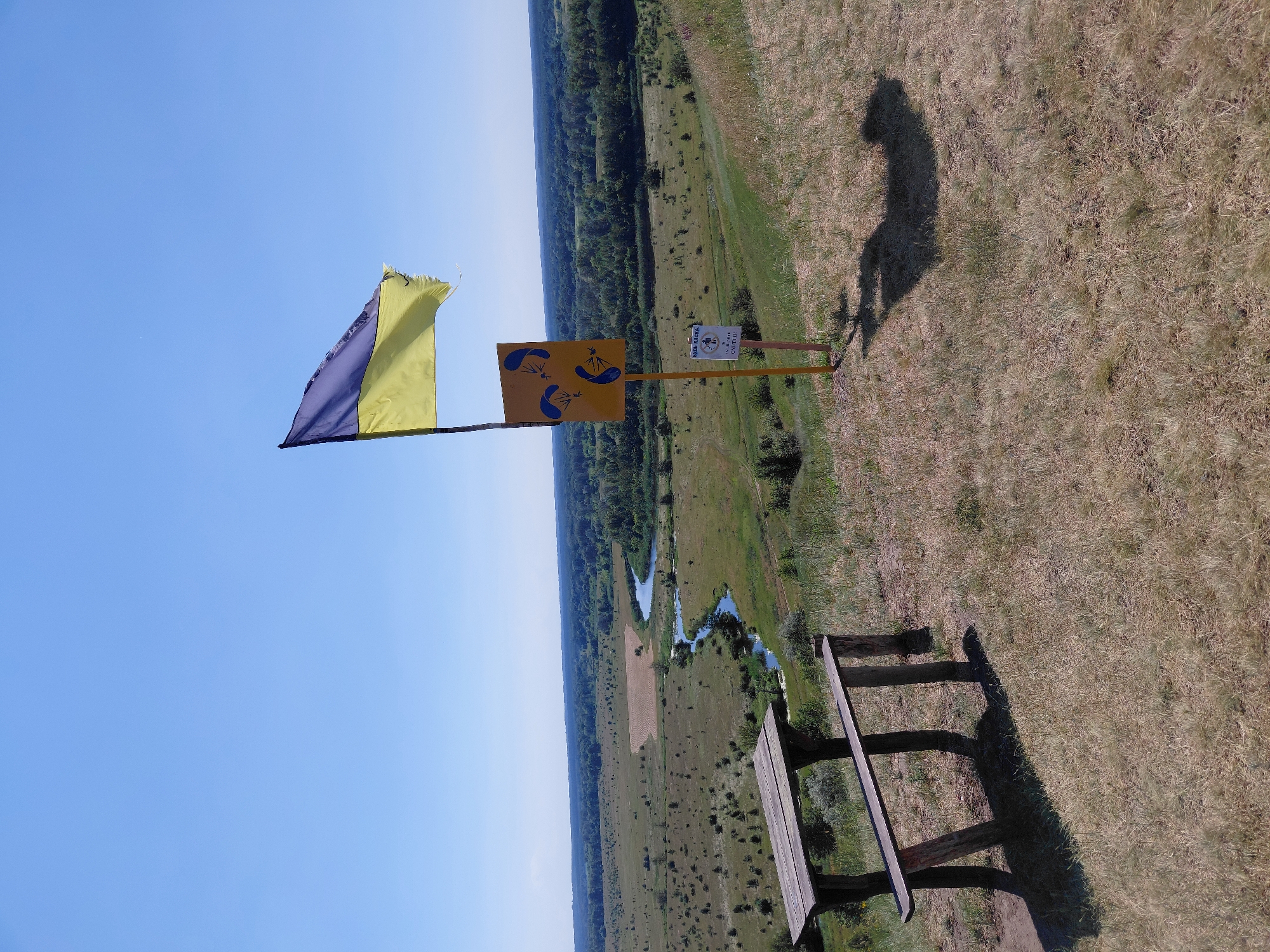 This image captures an outdoor scene under a clear blue sky, likely during the daytime on a sunny day. The orientation of the image is rotated 90 degrees counter-clockwise.
The main subjects include a flag with purple and yellow sections, attached to a pole. Below the flag, another pole supports a square sign with a yellow background, featuring three blue parachute silhouettes. A wooden picnic bench is positioned in the foreground, made of rough-hewn logs and planks, casting a long shadow on the dry, grassy ground.
In the background, a winding river or stream flows through a landscape of green fields and trees. A path or track is visible in the field. A person is standing near the signpost, partially obscured, and their silhouette is also visible on the ground, appearing to be a shadow cast by a person with arms raised.
A small sign is attached to the same pole as the yellow parachute sign, with visible text. One part of the sign appears to read "ПУНКТ ВІДЛЕТУ" (which translates to "Departure Point" in Ukrainian), and another section, below a clock symbol, says "СИРЕНА" (which translates to "Siren"). This suggests the location is a launch point, possibly for paragliding or similar activities, with an indication of an alert system.
The overall setting is rural and natural, with evidence of recreational activity. The presence of the flag, sign, and picnic bench suggests a designated area for visitors or participants. The time of day is likely late morning or afternoon, given the length and angle of the shadows. The weather appears to be fair and warm.