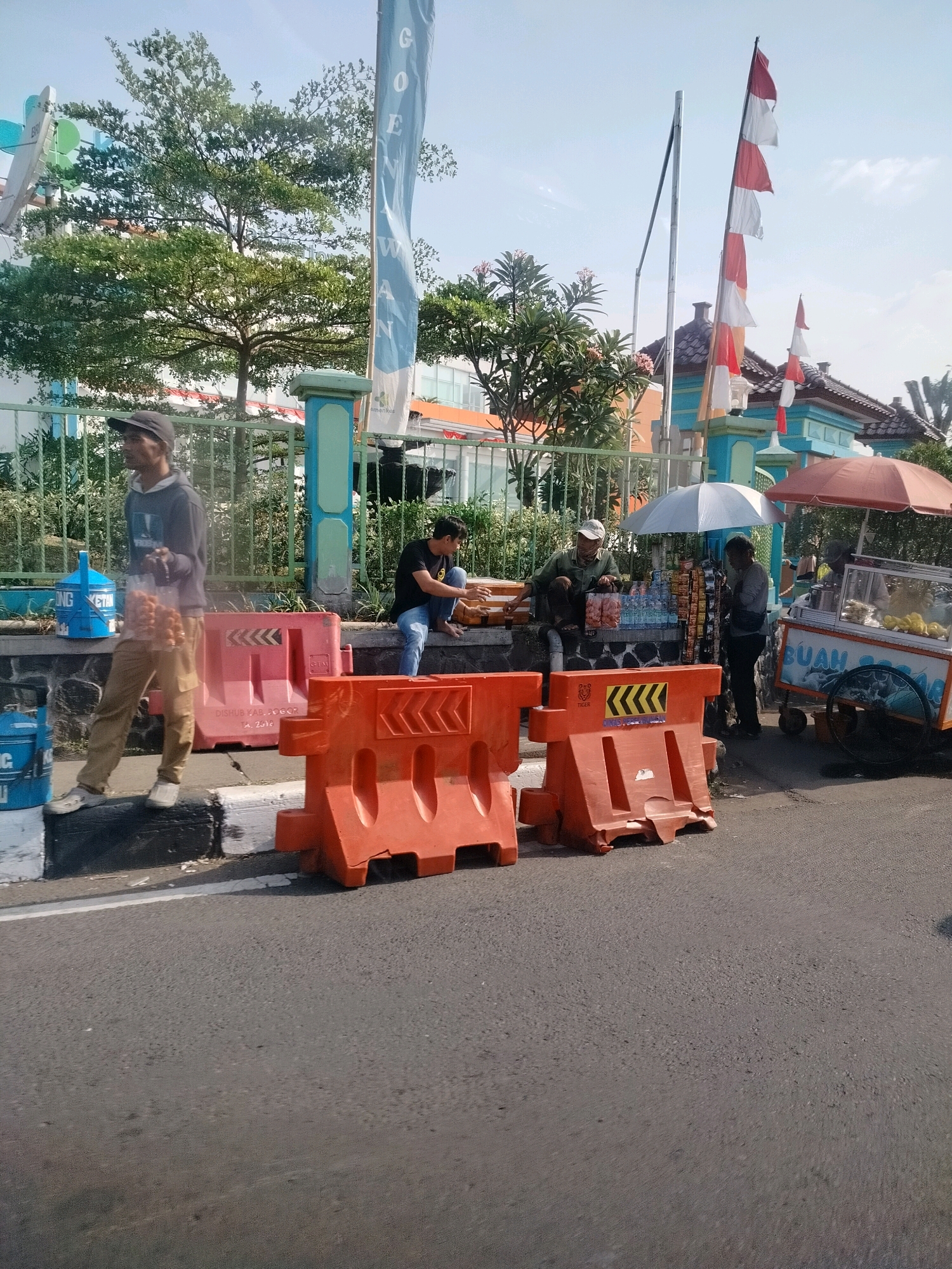 The image depicts a street scene in Cisarua, Indonesia, on a sunny day. In the foreground, a man wearing a dark hoodie, tan pants, and a baseball cap walks to the left, holding a plastic bag containing small, round items, presumably food. He is standing on a sidewalk next to several large, orange traffic barriers. To his right, a blue water dispenser with white lettering is visible. Further down the sidewalk, to the right of the traffic barriers, two men are seated on a stone ledge. One man, wearing a dark t-shirt and blue jeans, is looking to the left. The other man is wearing a lighter-colored shirt and a white cap, and he appears to be interacting with items on the ledge. Behind the seated men, a street vendor's cart is visible. The cart has an umbrella overhead and displays a variety of items, including what appears to be fruit. Another vendor, wearing a gray long-sleeved shirt and dark pants, stands near the cart. In the background, a fence runs along the edge of the property, and behind it are trees, buildings, and a flagpole with Indonesian flags. The overall environment suggests a commercial or public area with pedestrian and vehicular traffic.