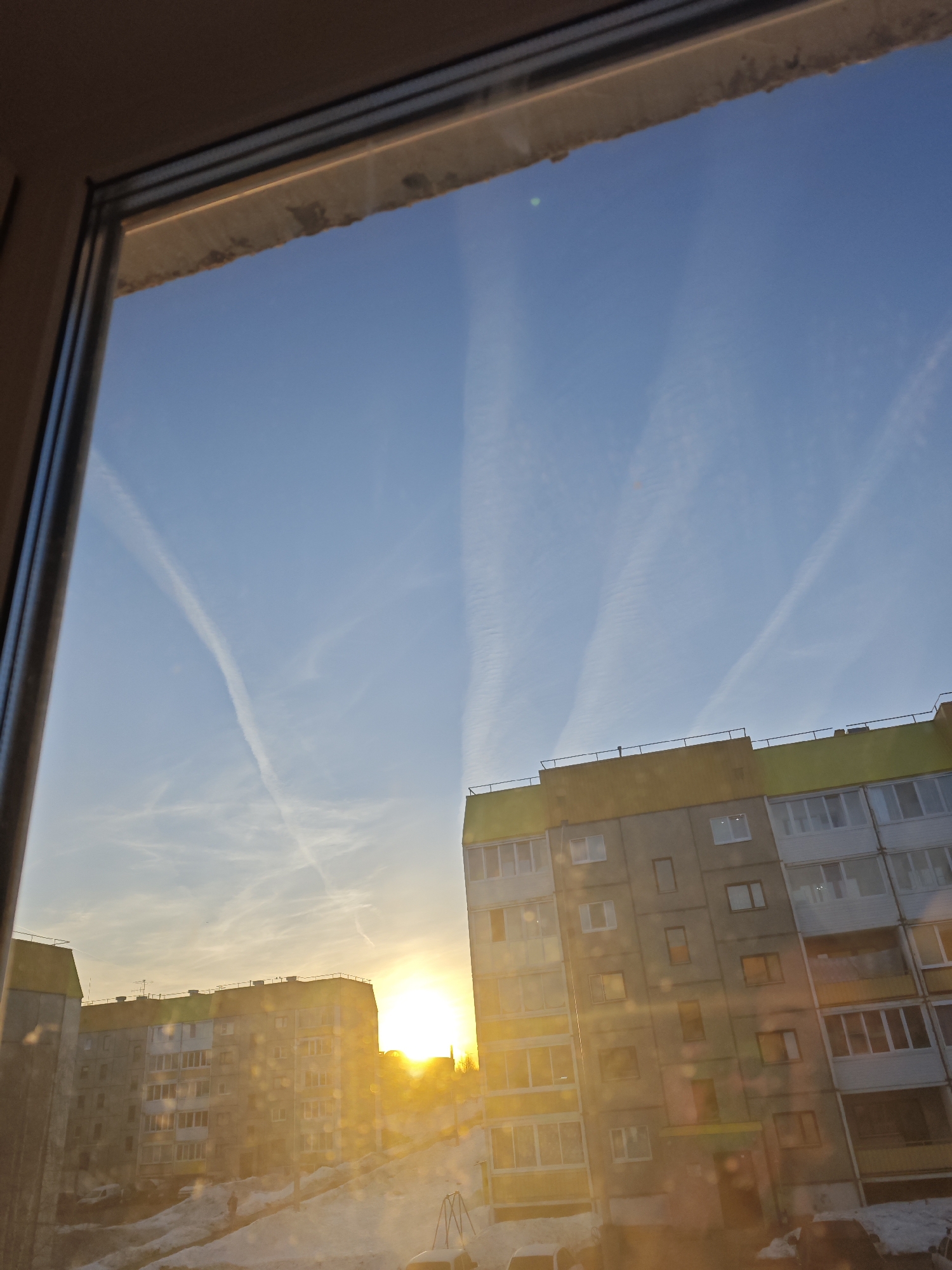 A low-angle shot through a window shows a view of apartment buildings in Markovskii, Russia. The buildings are predominantly gray with yellow and green accents on the upper levels, and the windows reflect the bright light of the setting sun. Streaks of condensation trails are visible in the clear blue sky. Some snow is present on the ground below. A window frame surrounds the scene, indicating the shot was taken from inside.