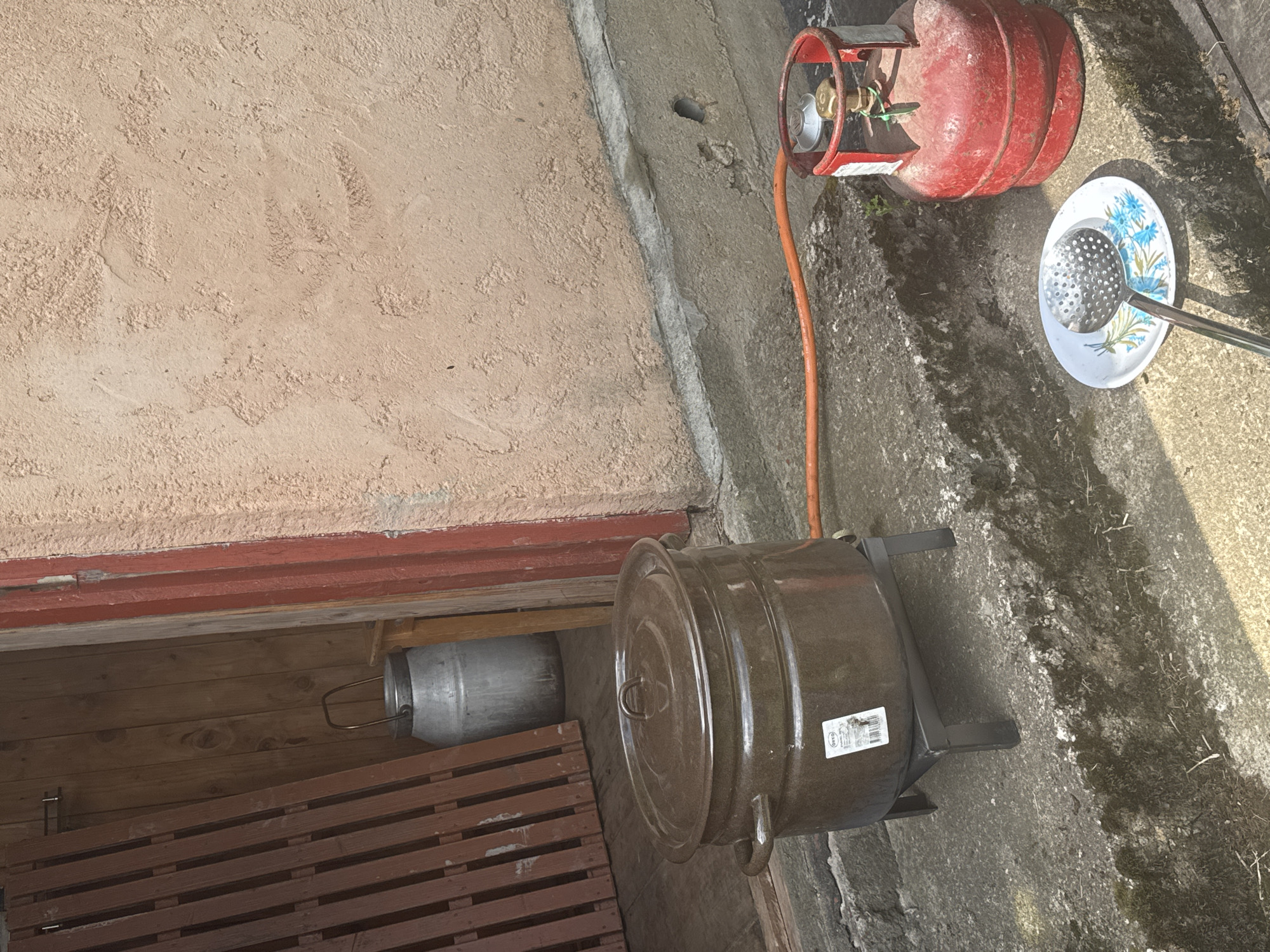 The image depicts an outdoor scene, possibly in the city of Rezekne, Latvia, where a large, dark-colored pot is set up on a black metal stand on concrete steps. An orange gas hose connects the pot to a red gas cylinder situated to the right. A small plate with a floral pattern holds a metal slotted spoon. In the background, an open doorway reveals wooden walls and a tall, metallic container. The overall environment suggests a rustic or utilitarian setting. There are no people present in the image.