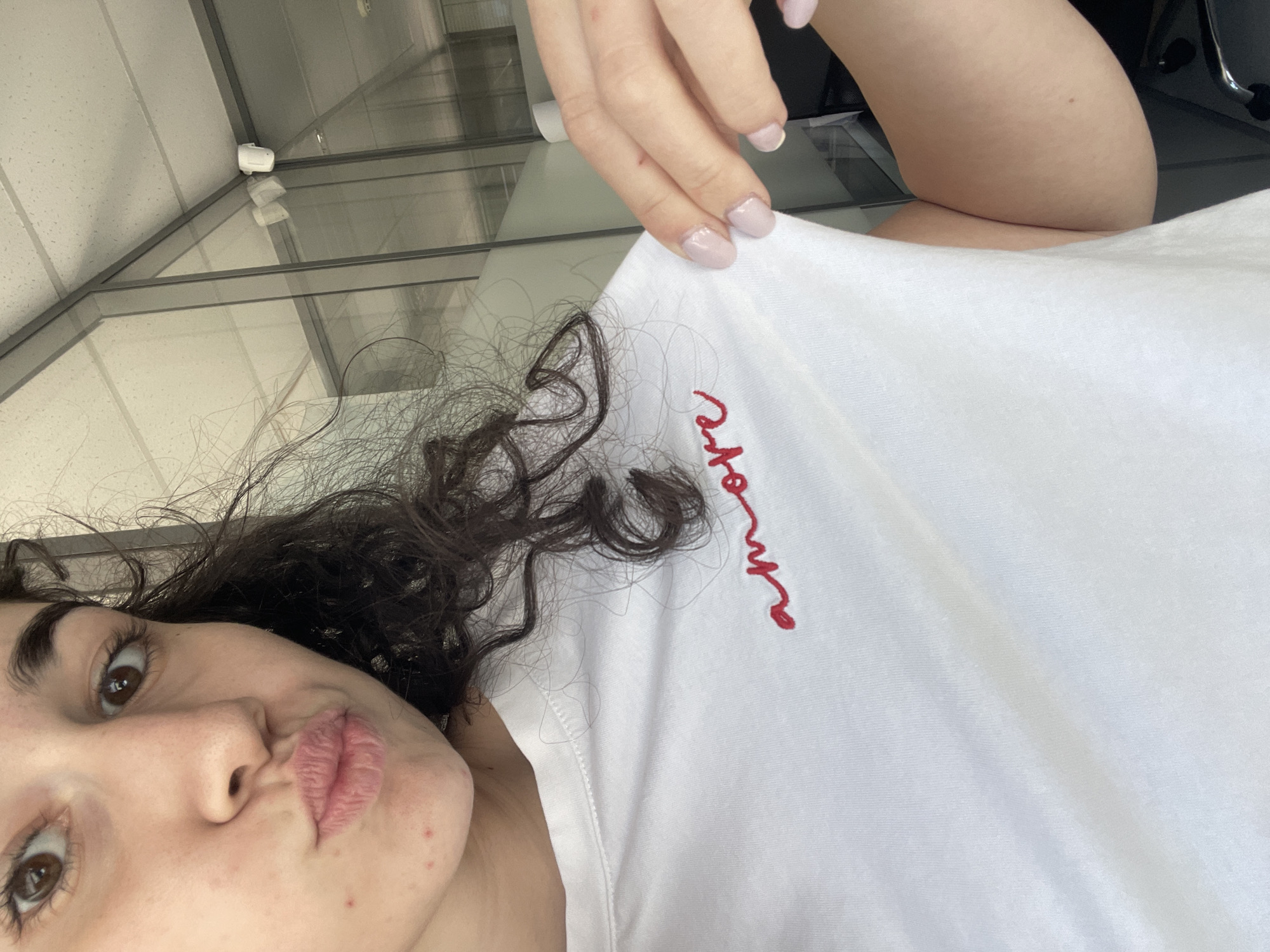 A woman with curly brown hair and light skin is taking a selfie. She is wearing a white t-shirt with the word "amore" embroidered in red on the upper chest. Her lips are pursed in a kissing expression. Her fingernails are painted a light pink color. The background appears to be a modern office space with a glass facade and visible ceiling fixtures. The photograph might have been taken indoors. There is no verifiable information to suggest the location is in Sofia, Bulgaria.