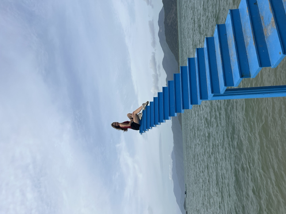 The image depicts a female subject seated on a blue staircase extending over water in Ninh Ích, Vietnam. The subject, with long brown hair, wears a dark red sleeveless top, black shorts, and light-colored sneakers. She is positioned sideways on an upper step of the staircase, with her left leg extended and her right knee bent, looking directly at the viewer with a smile. The staircase, constructed of bright blue material, rises diagonally from the lower right foreground towards the upper left, with its base partially submerged in or immediately adjacent to the water. The water body, appearing as a bay or sea, shows small ripples. In the distant left background, small dark structures are visible on the water. A range of dark green mountains or hills lines the horizon, separating the water from a light blue sky with scattered white and grey clouds.