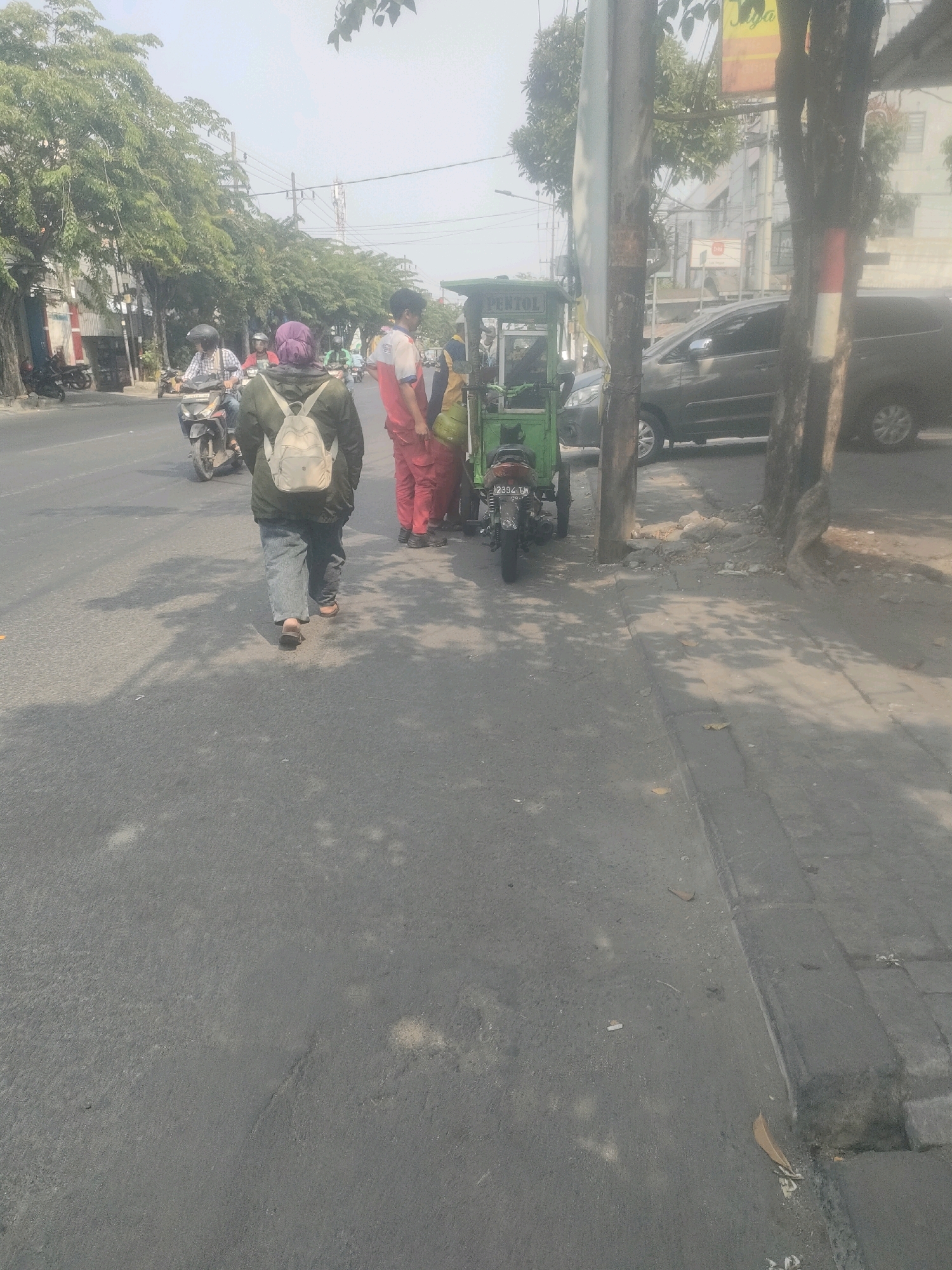In Surabaya, Indonesia, a street scene unfolds under a hazy sky. A person wearing a purple headscarf and a green jacket with a light-colored backpack walks away from the camera on a gray asphalt road. To their right, a green mobile cart, labeled "PENTOL," is parked near a utility pole. Two individuals stand near the cart; one in a red and white uniform and another in red pants. A motorcycle with license plate number B 2394 TW is parked behind the cart. In the background, vehicles, including a dark-colored minivan, are visible on the road and a sidewalk runs alongside the street. Trees with dense foliage line the left side of the road, and buildings are visible in the distance.