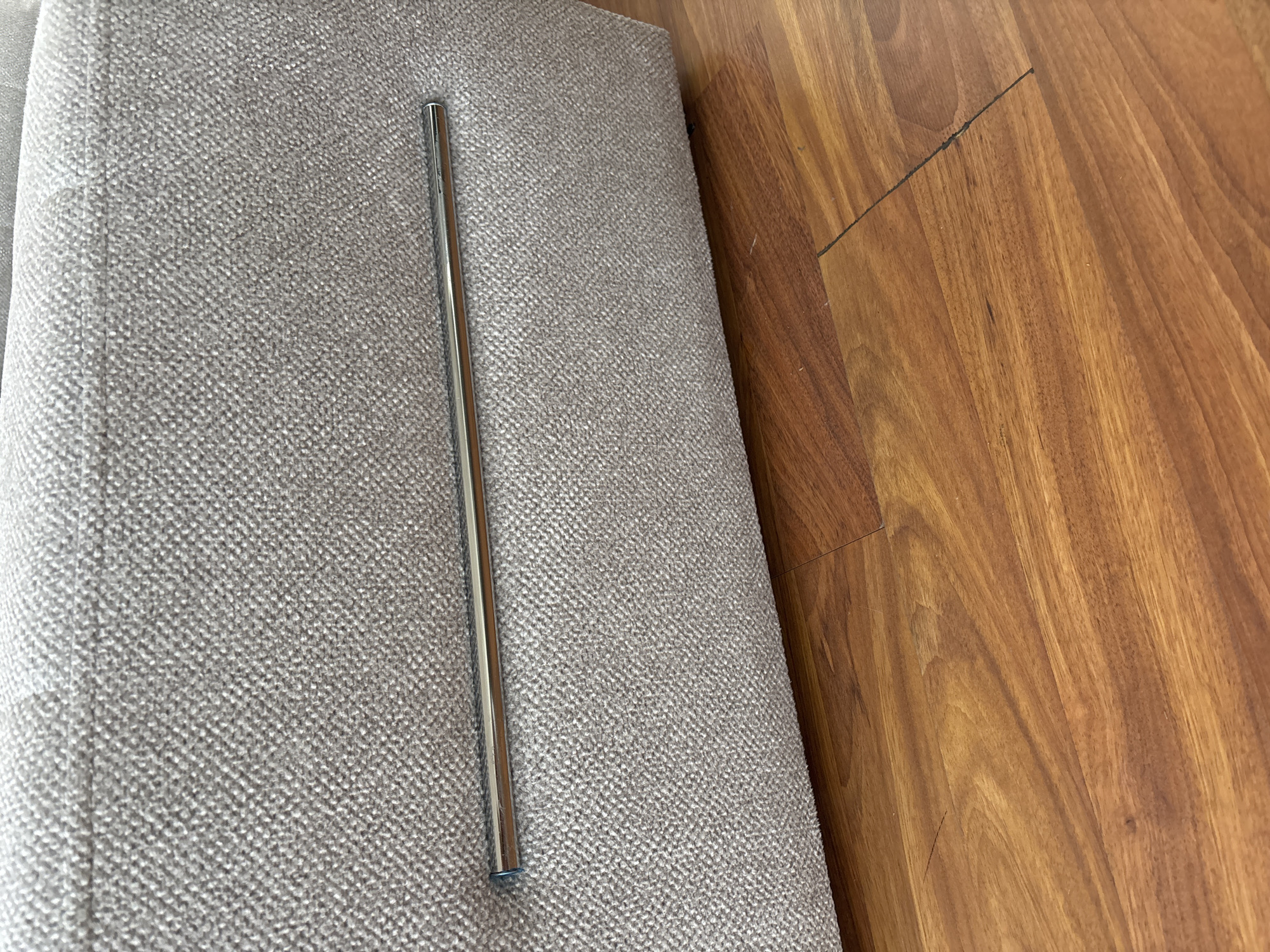A close-up shot shows a portion of a beige upholstered sofa with a metal bar across its front. The sofa is placed on a wooden floor with a visible grain pattern. The image is indoors and focuses on the texture of the fabric and the sheen of the metal bar. There are no people present in the image. The location is not specified within a city.