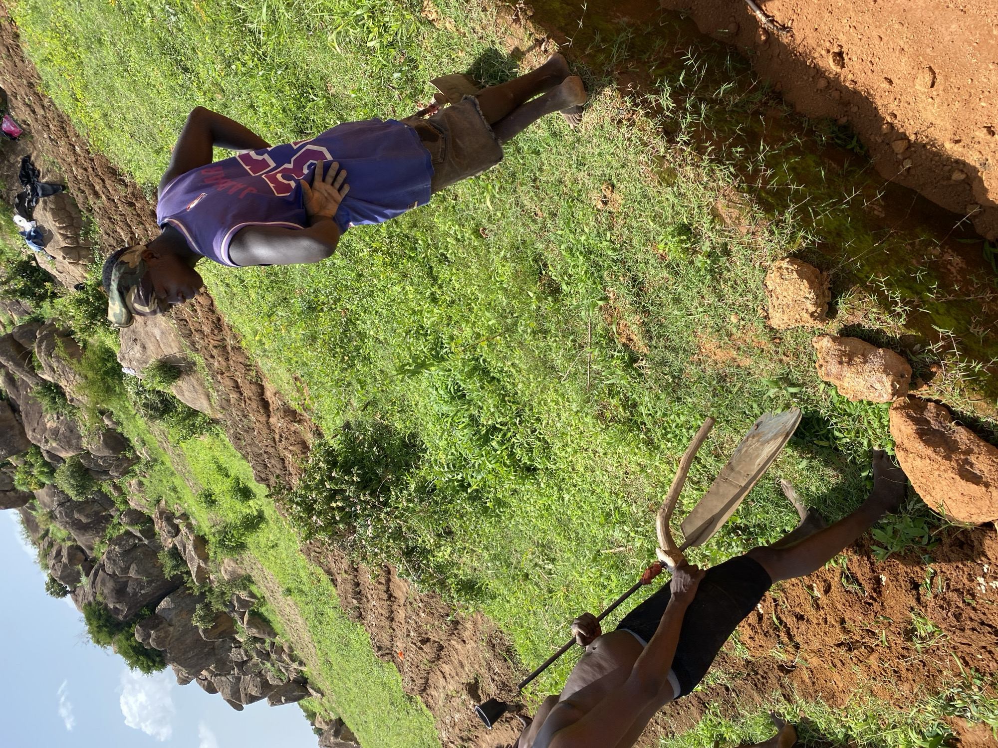 In the city of Bukuru, Nigeria, two individuals are engaged in manual labor on a steep, grassy hillside under a bright sky. One man, wearing a purple basketball jersey with the number 35 and a camouflage headband, stands with his hand on his hip, observing. The other man, shirtless and wearing dark shorts, is actively digging with a traditional hoe, a wooden tool with a flat, wide blade. The terrain is characterized by rocky outcrops and patches of both barren, reddish soil and lush green vegetation. The men appear to be working in a trench or ditch that has been dug into the hillside, with piles of excavated earth present. The overall scene depicts agricultural or construction work being carried out in a rugged, natural environment.