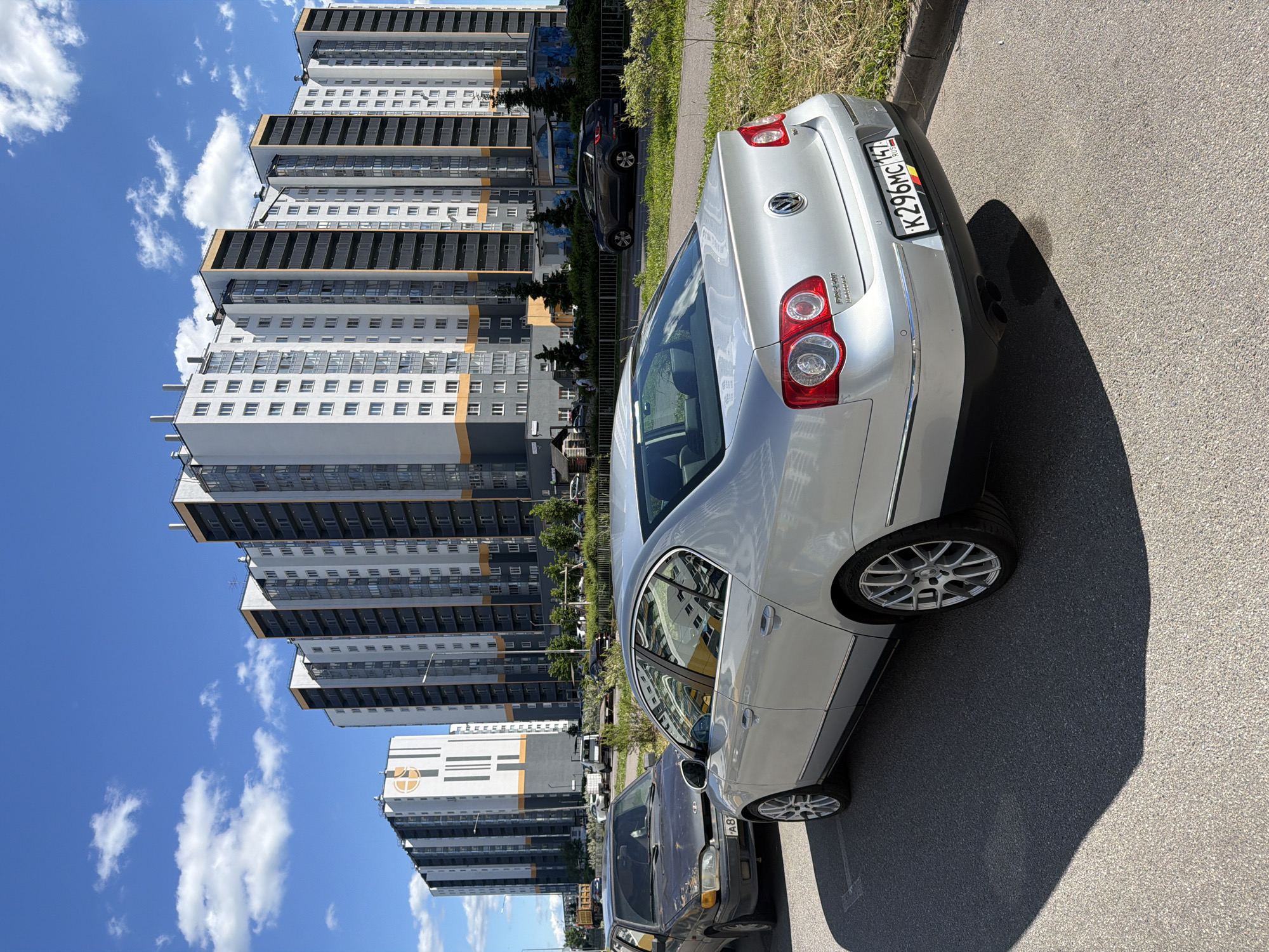 A vertical, eye-level shot captures a sunlit scene of parked cars and modern apartment buildings under a bright blue sky with scattered white clouds. The main subject is a silver Volkswagen sedan parked on an asphalt road, its rear prominently displayed. The car's license plate is visible, bearing the text "K296MC147". To the left of the car, a series of tall, multi-story apartment buildings dominates the background. These buildings feature a repeating pattern of white and gray facades with numerous windows, accented by dark horizontal strips and some orange detailing. The buildings appear modern and residential. Further back, beyond the apartment buildings, a darker colored car is also parked, and glimpses of trees and greenery can be seen. The scene suggests a daytime setting with clear weather, likely in a suburban or urban residential area. The overall impression is one of a quiet, everyday moment captured outdoors.