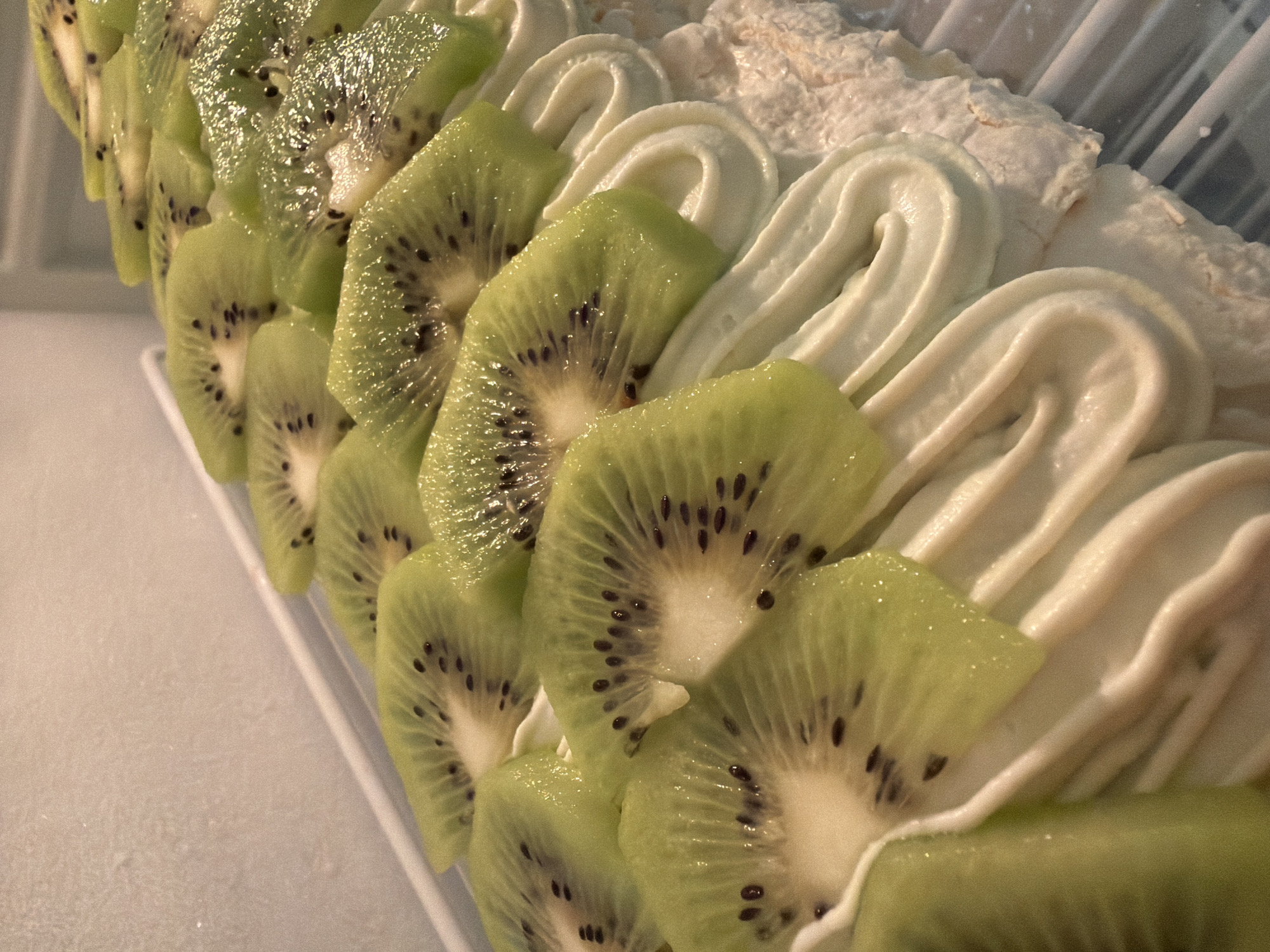 A close-up shot of a dessert, likely photographed in Tel-Aviv, Israel, given the prompt. The dessert features neatly arranged kiwi slices and piped white cream. The kiwi slices are bright green with distinct black seeds. The white cream appears smooth and thick, and the dessert rests on a white or light-colored base. The light suggests an indoor setting. No people are visible in the frame.