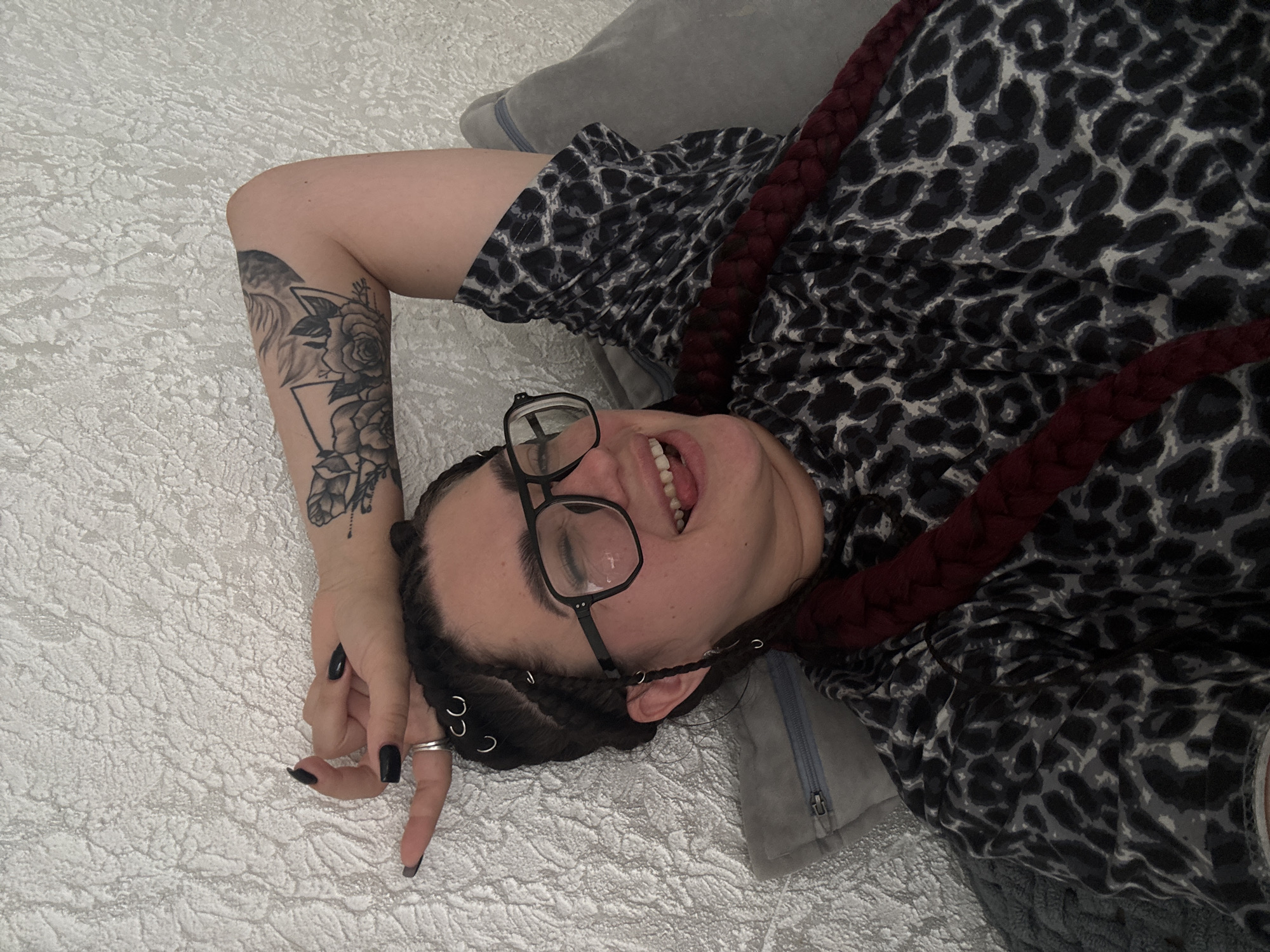 An overhead shot shows a woman lying on a textured, off-white surface, possibly a wall or headboard. She is wearing glasses, a black and grey leopard-print top, and has her left arm raised behind her head. Her hair is styled in braids with silver rings woven in. Her right arm is bent, with her hand near her face. She is sticking her tongue out and her eyes are closed. There is a grey pillow behind her head and a dark grey blanket beneath her to the left. A dark red braid is visible across her chest.