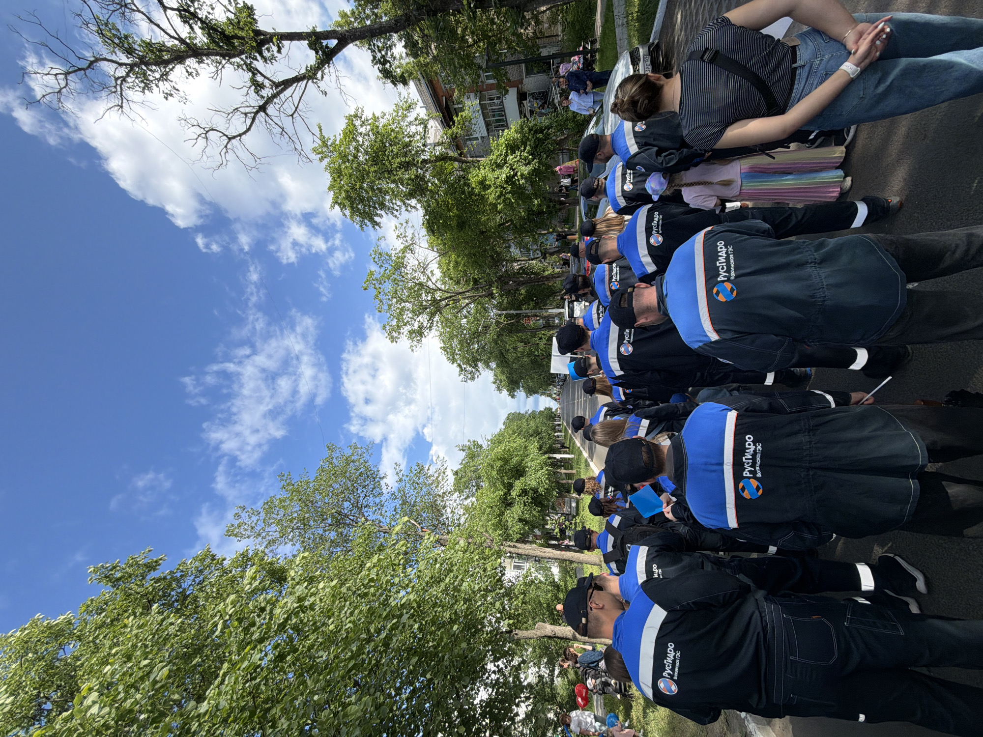 The image depicts a group of people, mostly men, wearing matching dark blue jackets with blue and orange accents and logos that read "Русгидро" and "Воткинская ГЭС" in Cyrillic. They are walking down a tree-lined street in the city of Chaikovsky, Russia. The sky is blue with scattered white clouds, and the sun appears to be shining. Several trees with full green foliage flank the street, and apartment buildings are visible in the background. Some individuals in the group are holding blue flags. The scene suggests a organized event or procession, possibly related to the energy company "RusHydro," which operates the Votkinsk Hydroelectric Power Station.