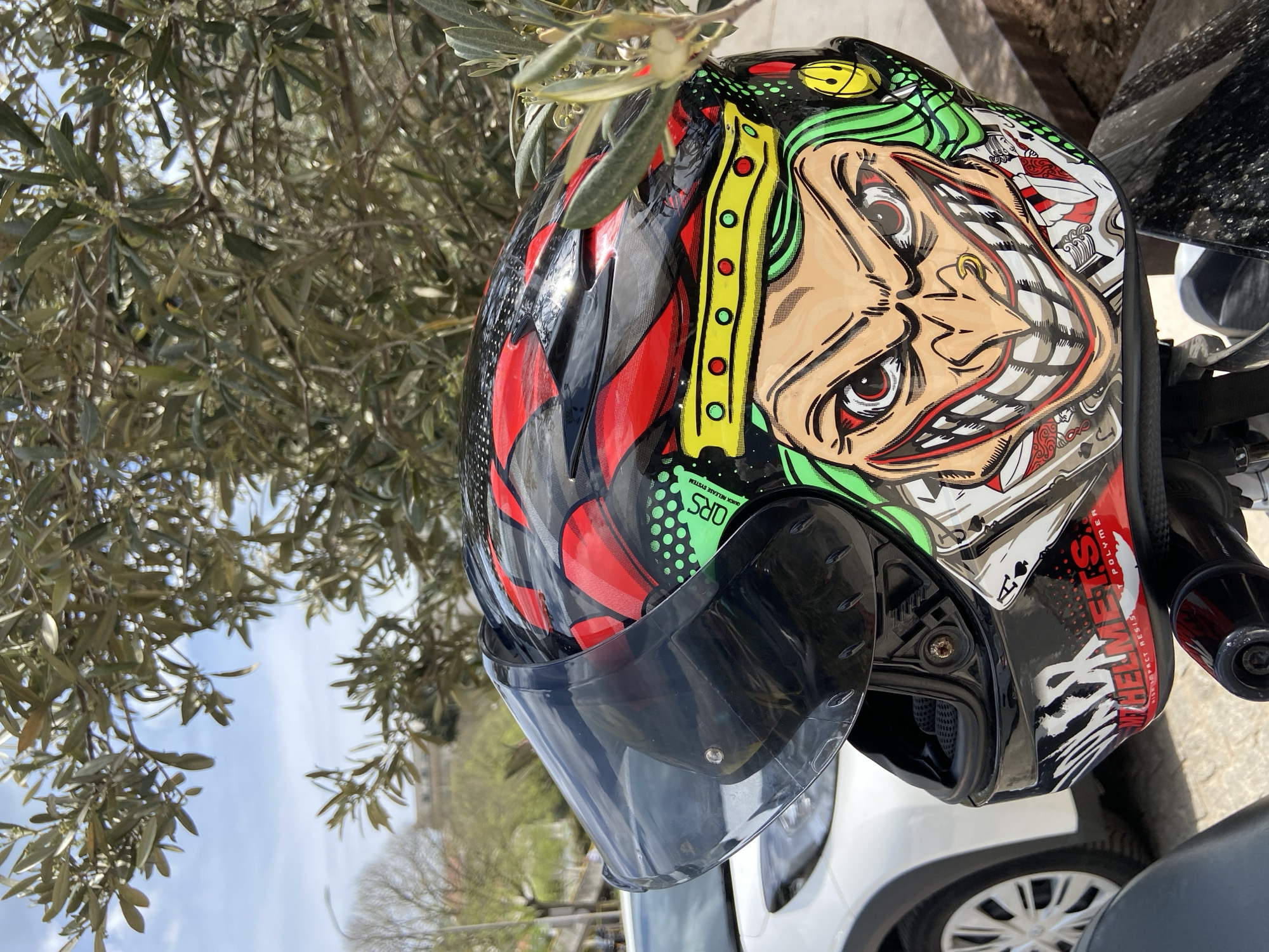 An angled, close-up shot captures a motorcycle helmet adorned with a vivid, detailed graphic design. The helmet, predominantly black, features a stylized, cartoonish face with a wide, exaggerated grin, large eyes, green hair, and a nose ring, reminiscent of a joker or clown. The design incorporates elements of playing cards, including an ace of spades, and includes red and black abstract or flame-like patterns. A dark, clear visor is partially raised on the helmet.
The helmet is positioned outdoors, likely resting on a motorcycle or scooter, with part of its handlebars visible on the right. In the background, numerous branches and leaves of an olive tree are prominent, with some small, dark olives visible among the foliage, suggesting a Mediterranean climate. Above and behind the tree, a clear blue sky with some soft clouds is visible. In the lower part of the image, the side and wheel of a white car are partially seen, suggesting a parked vehicle on a street or in a parking area. Distant, blurred trees and buildings hint at an urban or suburban setting.
The scene is brightly lit by natural sunlight, indicating daytime. Visible text on the helmet includes "QUICK RELEASE SYSTEM" and "MY HELMETS HIGH IMPACT POLYMER". The overall impression is a vibrant object set against a natural and urban backdrop, consistent with the Budva, Montenegro context.