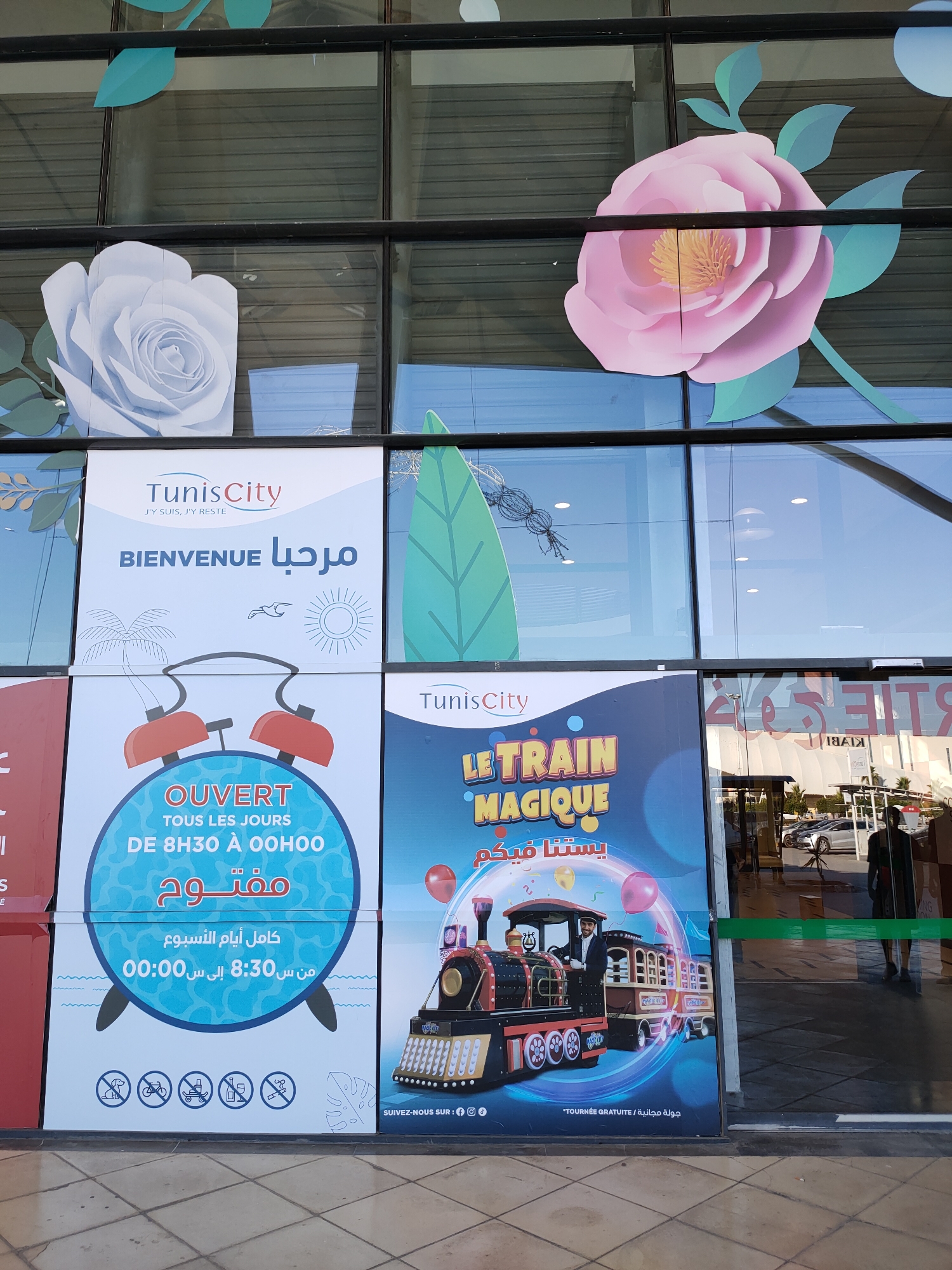 The image shows the exterior of a building with large glass windows, likely a shopping mall or public venue. The windows are decorated with large, stylized paper flowers in white and pink, and green leaf cutouts.
On the left side, there is a large poster with the text "TunisCity" and "BIENVENUE" in large blue letters, accompanied by Arabic script. Below this, there is an illustration of an alarm clock with the text "OUVERT TOUS LES JOURS DE 8H30 À 00H00" and Arabic text stating "مفتوح كامل أيام الأسبوع من س 8:30 إلى س 00:00". At the bottom of this poster, there are icons depicting prohibited activities: a dog, a bicycle, a stroller, a bottle, and a person smoking.
In the center, there is a poster featuring a colorful illustration of a toy train with the text "LE TRAIN MAGIQUE" in yellow letters and Arabic text below it. Balloons are depicted around the train. At the bottom of this poster, it says "SUIVEZ-NOUS SUR:" followed by social media icons and "TOURNÉE GRATUITE / جولة مجانية".
To the right of the posters, there is a glass entrance with a green band across it. Reflections in the glass show cars and people inside a brightly lit space. The ground in front of the building is paved with square tiles. The overall impression is of an advertisement for a shopping center in Tunis City, Tunisia, highlighting its hours of operation and a children's train attraction.