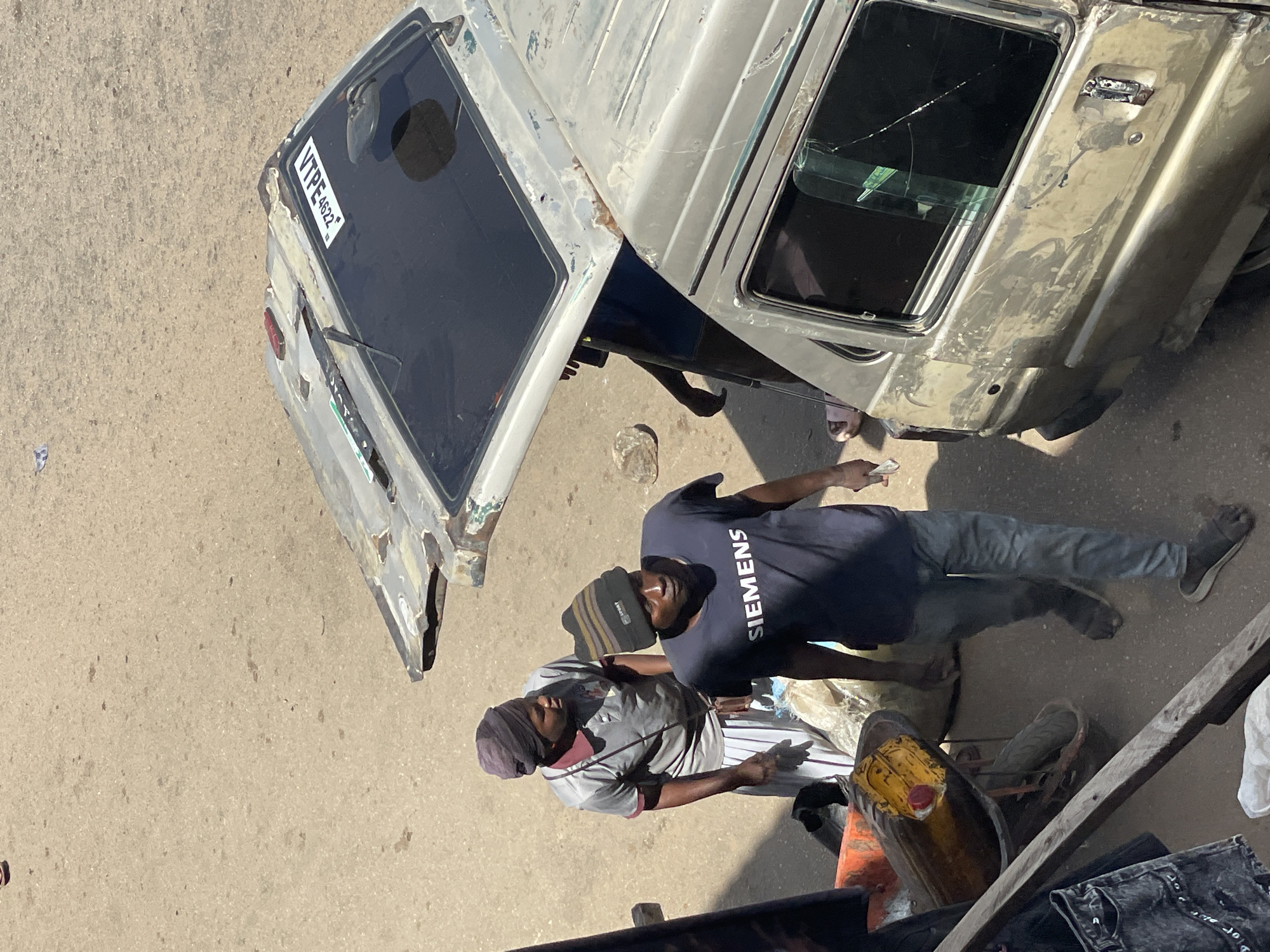 The image shows an outdoor scene, presumably in Bukuru, Nigeria, given the license plate "VTPE 4622" of the vehicle present. Two men are standing near a dilapidated, silver/gray van or truck. One man is wearing a dark blue t-shirt with "SIEMENS" printed on it, grey jeans, and black shoes. He also has a dark-colored beanie. The other man is wearing a grey shirt with dark stripes, light-colored pants, and a dark head covering. A yellow jerrycan sits nearby. The vehicle has apparent damage, including a cracked windshield and rust. The road surface is concrete.