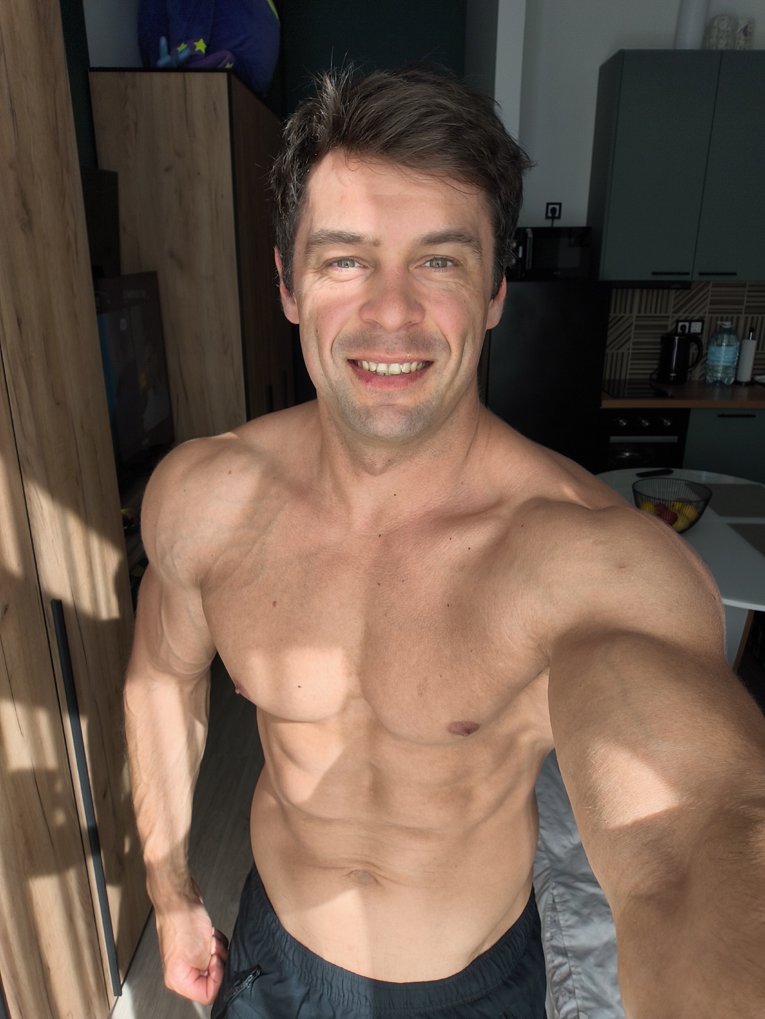 A shirtless man is taking a selfie indoors. He is wearing black shorts. The background includes wooden cabinets, a black refrigerator, kitchen cabinets in a dark color, and a table with a fruit bowl. The location is not specified in the image and thus cannot be identified as Moscow, Russia.