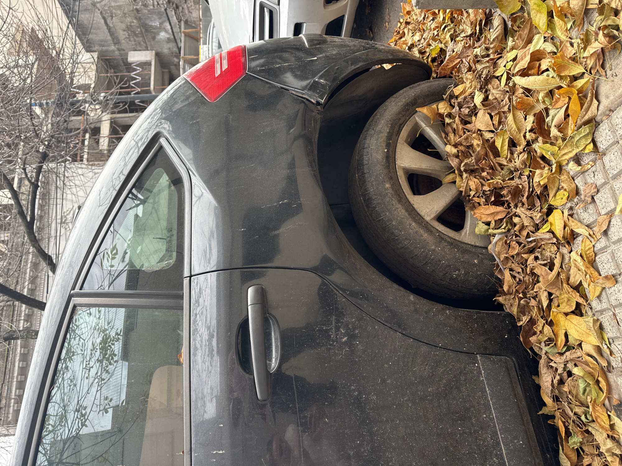 A black car is parked on a sidewalk in Buenos Aires, Argentina, surrounded by fallen leaves. The car appears dirty and may be damaged. A building is visible in the background, suggesting an urban setting.