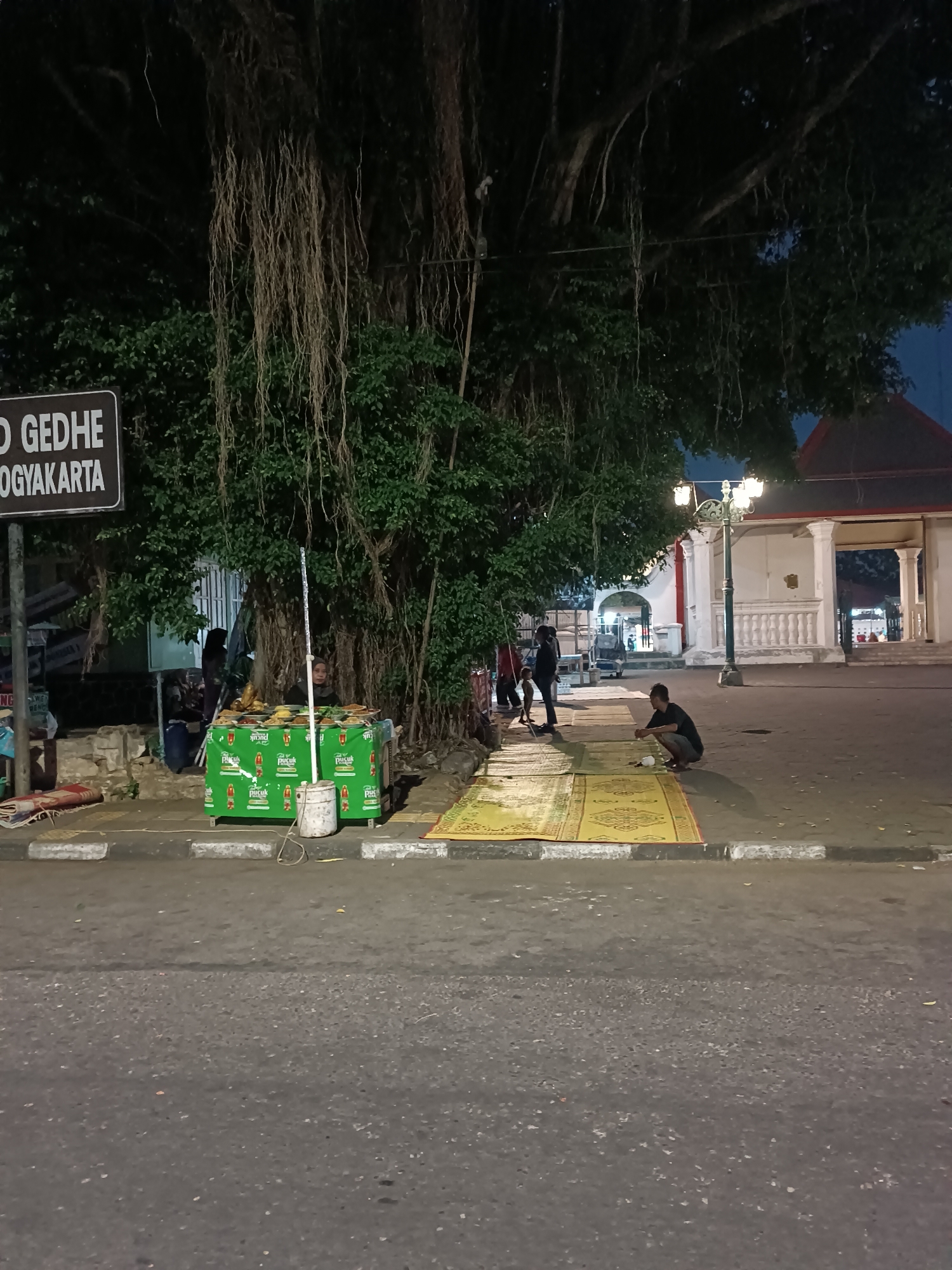 The image depicts a street scene at night in Yogyakarta, Indonesia, likely near a public square or heritage site, suggested by the sign reading "GEDHE YOGYAKARTA." A large banyan tree with hanging aerial roots dominates the left side of the frame, casting shadows. In the foreground, a street vendor's cart is set up, displaying food items under a green canopy. Several people are visible: a vendor behind the cart, a few individuals standing and interacting near the cart, and a man crouching on a yellow mat in the middle of the paved area. The man is facing away from the camera and appears to be engaged in an activity on the mat, which is spread out on the ground. A street lamp with multiple bulbs illuminates the background, where a white building with arched openings and columns is visible, possibly part of an architectural complex. The street is paved with asphalt, and a sidewalk with a white curb separates it from the area where the vendor and people are gathered. The overall atmosphere is dimly lit, typical of a street at night.