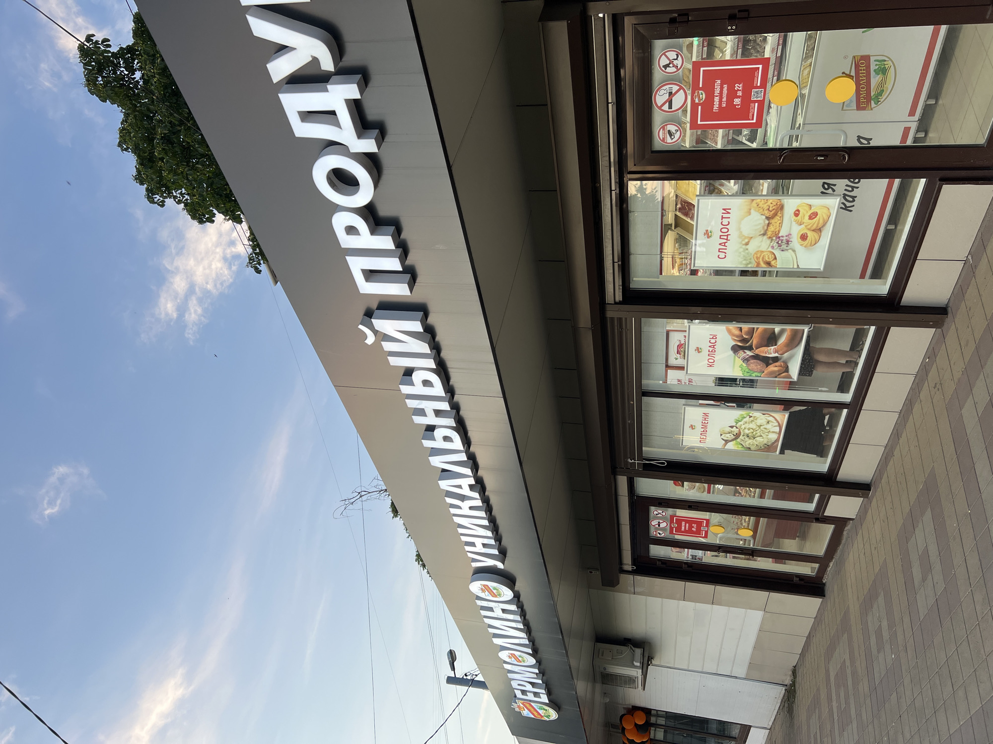 The image shows a storefront in Krasnodar, Russia, for a business named "ЕРМОЛИНО" (ER MOLINO), which appears to sell food products. The storefront has a large, illuminated sign across the top that reads "ЕРМОЛИНО УНИКАЛЬНЫЙ ПРОДУКТ" (ER MOLINO UNIQUE PRODUCT). The building has a modern facade with large glass windows displaying various food items. On the windows, there are posters advertising "ПЕЛЬМЕНИ" (pelmeni/dumplings) and "СЛАДОСТИ" (sweets). Inside, refrigerated display cases showcase products. There are also several warning signs on the door, including one indicating no smoking and another showing prohibited activities. The pavement in front of the store is paved with decorative tiles. The sky above is blue with some scattered clouds. There are no people clearly visible in the image, though legs of individuals are partially visible through the windows. The overall impression is that of a retail establishment.