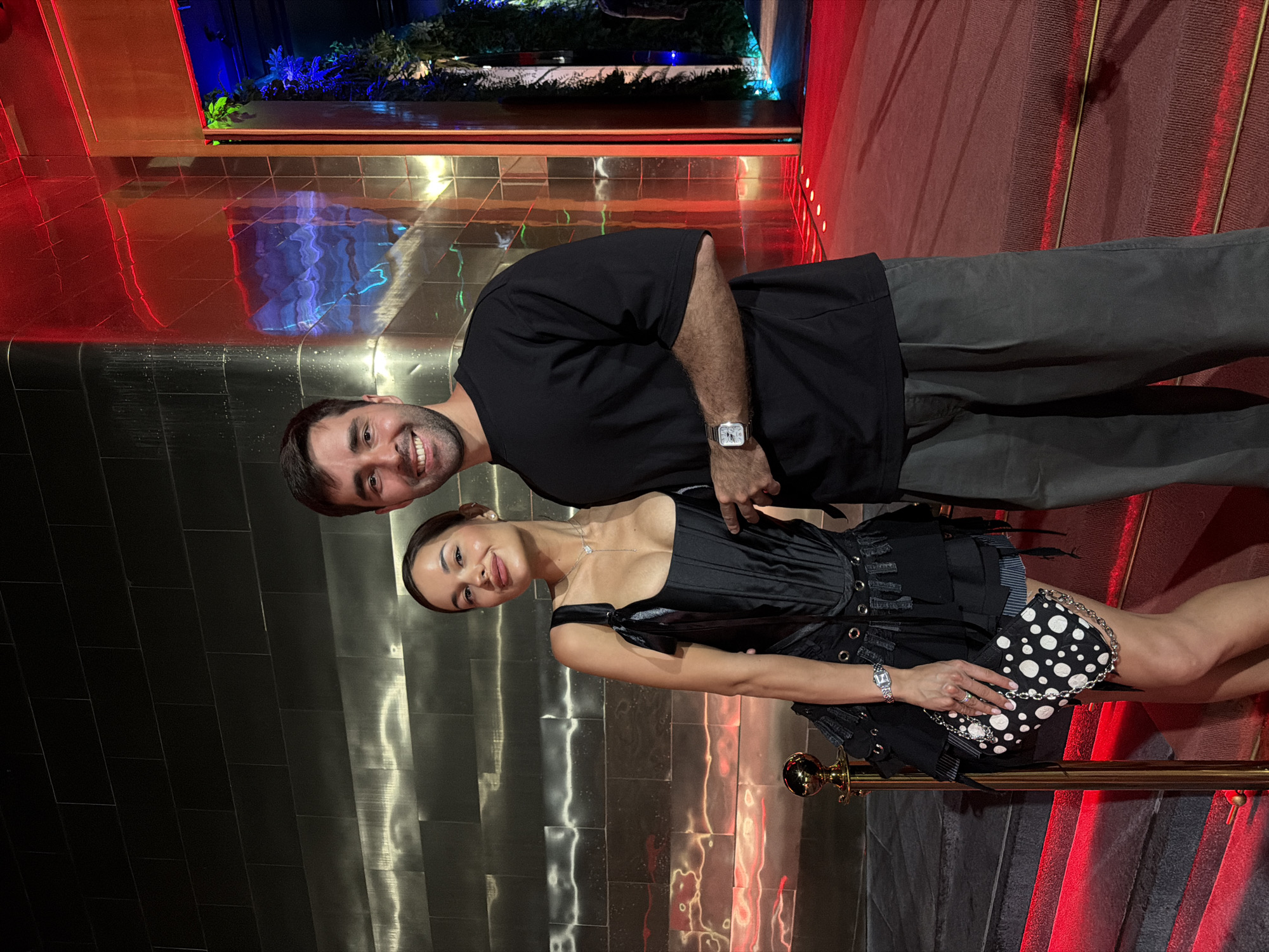 The image shows a man and a woman posing for a photo in what appears to be a high-end venue. The woman is on the left, wearing a black corset top, short skirt, and holding a black and white polka-dot purse. She has dark hair pulled back and is wearing a necklace and a watch. The man stands to her right, wearing a black t-shirt and gray pants. He is also wearing a watch. They are both smiling.
The background features a wall covered in metallic tiles and a section with reddish-brown tones that might indicate steps or an elevated area. There is also a lit aquarium-like display above, visible in the upper portion of the image.
The location is Choeng Thale, Thailand.