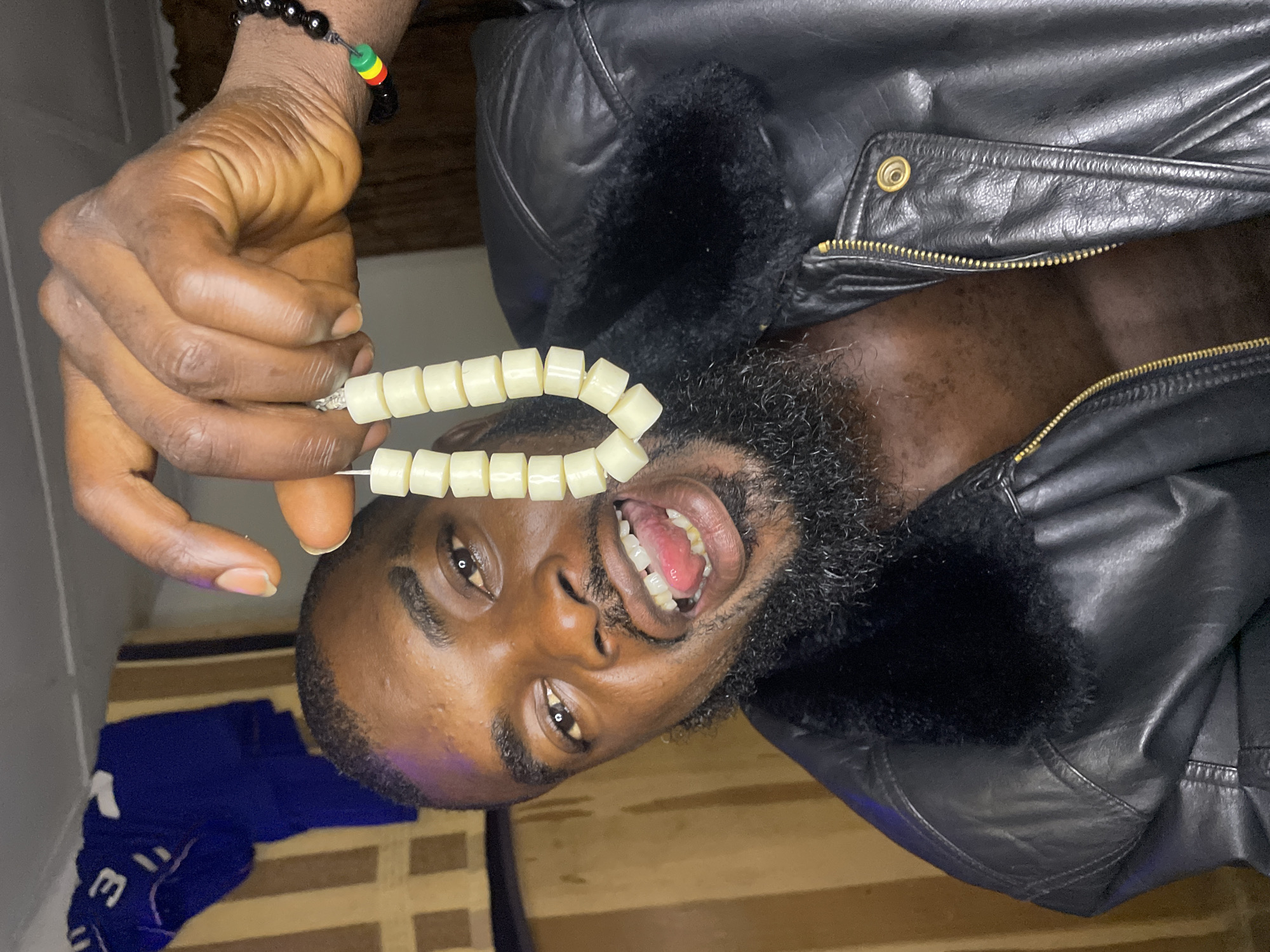 An adult male with dark skin, a black beard, and short dark hair is captured from the chest up in an indoor setting. He looks directly at the camera with a wide-open mouth, tongue slightly extended, and eyes open. He wears an unzipped black leather jacket featuring a black faux fur collar, revealing his bare chest. On his left wrist, a beaded bracelet with black beads and a segment of red, yellow, and green beads is visible. His right hand holds a string of uniformly shaped, light beige, rectangular beads, suspended in front of his face. The background consists of a draped blue fabric object and a vertical striped pattern in shades of beige and brown. There are no visual elements in the image to identify the specific location as Jos, Nigeria.