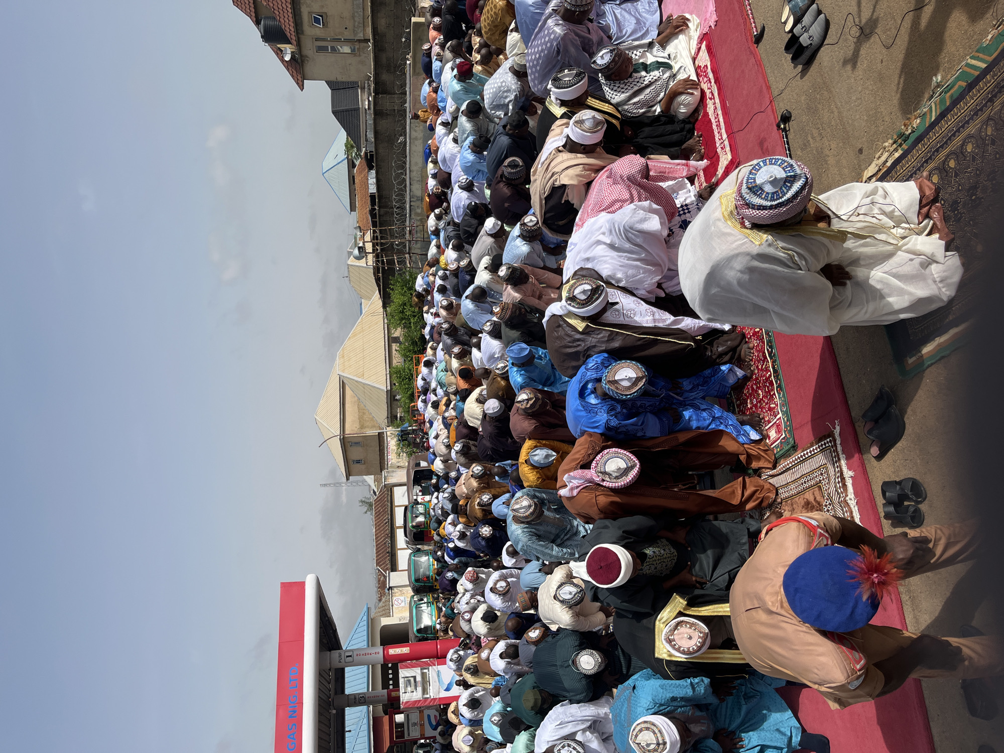 A large group of Muslim men are performing prayers outdoors on a red rug, near a gas station labeled "GAS NIG. LTD." The men are dressed in traditional Muslim clothing, including various styles of robes and hats. The scene appears to be in a densely built-up area with buildings visible in the background. The image is likely taken in Jos, Nigeria.