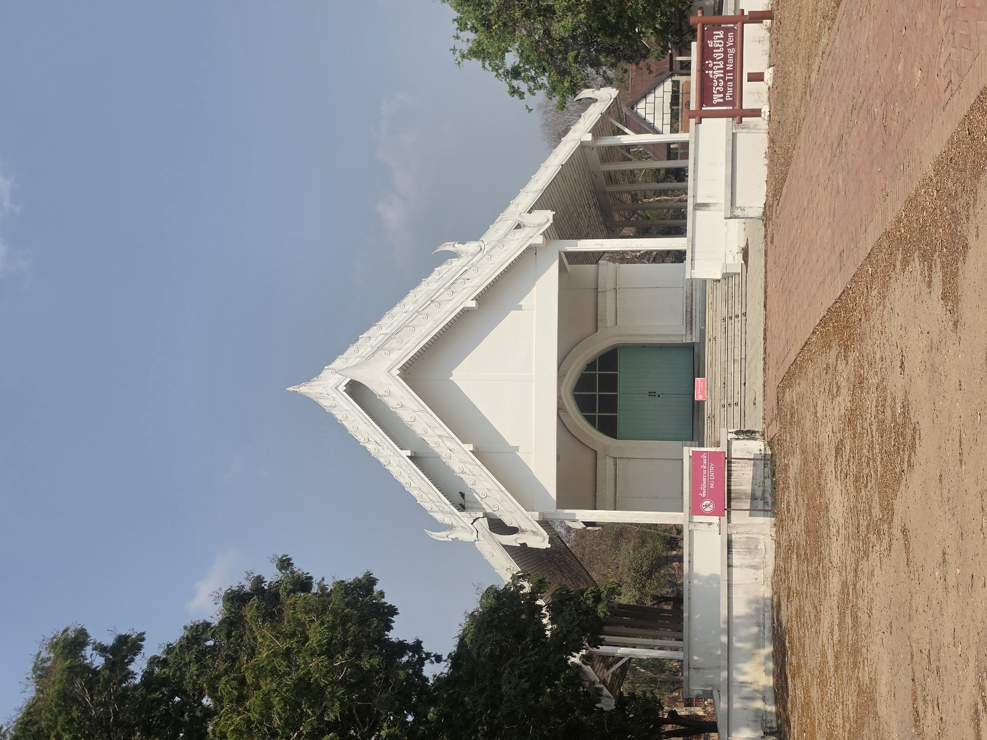 The image shows Phra Thinang Yen, a white building with a light green door in Phra Nakhon Si Ayutthaya City Municipality, Thailand. The building has a triangular roof with ornate details. There are trees on the left, and a brick path to the right. Signs indicate that entry is restricted. The sky is blue with some clouds.