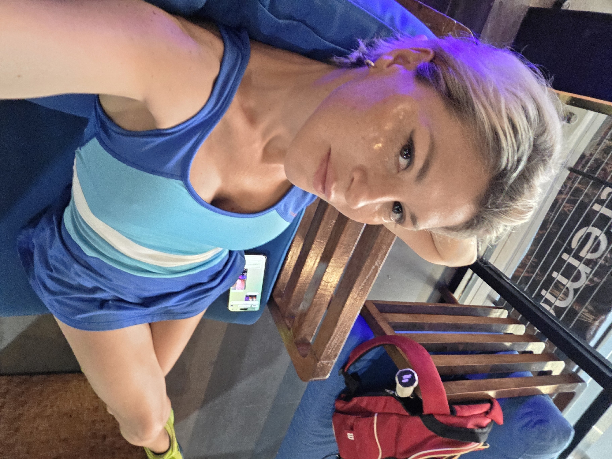 A blonde woman with a blue athletic top lies on a blue couch, her head resting on her arm. She has glitter makeup on her cheeks and eyes. A red backpack and a mobile phone are visible on the couch next to her. The setting appears to be an indoor lounge area with wooden chairs and blue seating. A banner with text is visible in the background. The scene is illuminated by a mix of ambient and purple lighting.
