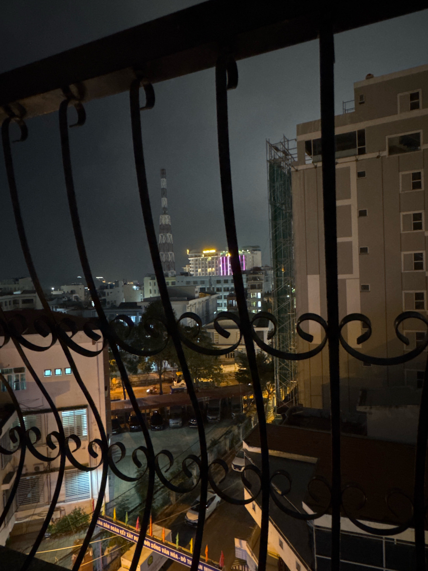 The image, captured at night, presents a view of an urban landscape from behind a decorative metal grate. In the foreground, the dark, ornate metal bars of a balcony or window frame dominate the lower portion of the image, obscuring parts of the scene beyond.
Beyond the grate, the cityscape unfolds. Several buildings of varying heights are visible. To the right, a large, modern, multi-story building with a beige facade and numerous windows stands prominently. Adjacent to it, a taller structure is under construction, encased in scaffolding and green netting. Further into the distance, other buildings are illuminated by interior lights or external lighting. A prominent radio or telecommunications tower with a lattice structure is visible in the middle ground, against a dark, overcast sky. Some buildings display colorful illuminated signage or lights, suggesting commercial activity or residential areas.
Below the buildings, the dark shapes of parked vehicles are discernible, and streetlights cast a yellow glow on the ground. Trees are scattered throughout the urban environment. The overall impression is that of a densely populated city at night, with ongoing development and typical urban infrastructure. The view suggests a location within the city of Nha Trang, Vietnam, given the style of buildings, the presence of the radio tower which could be a landmark, and the general urban context.