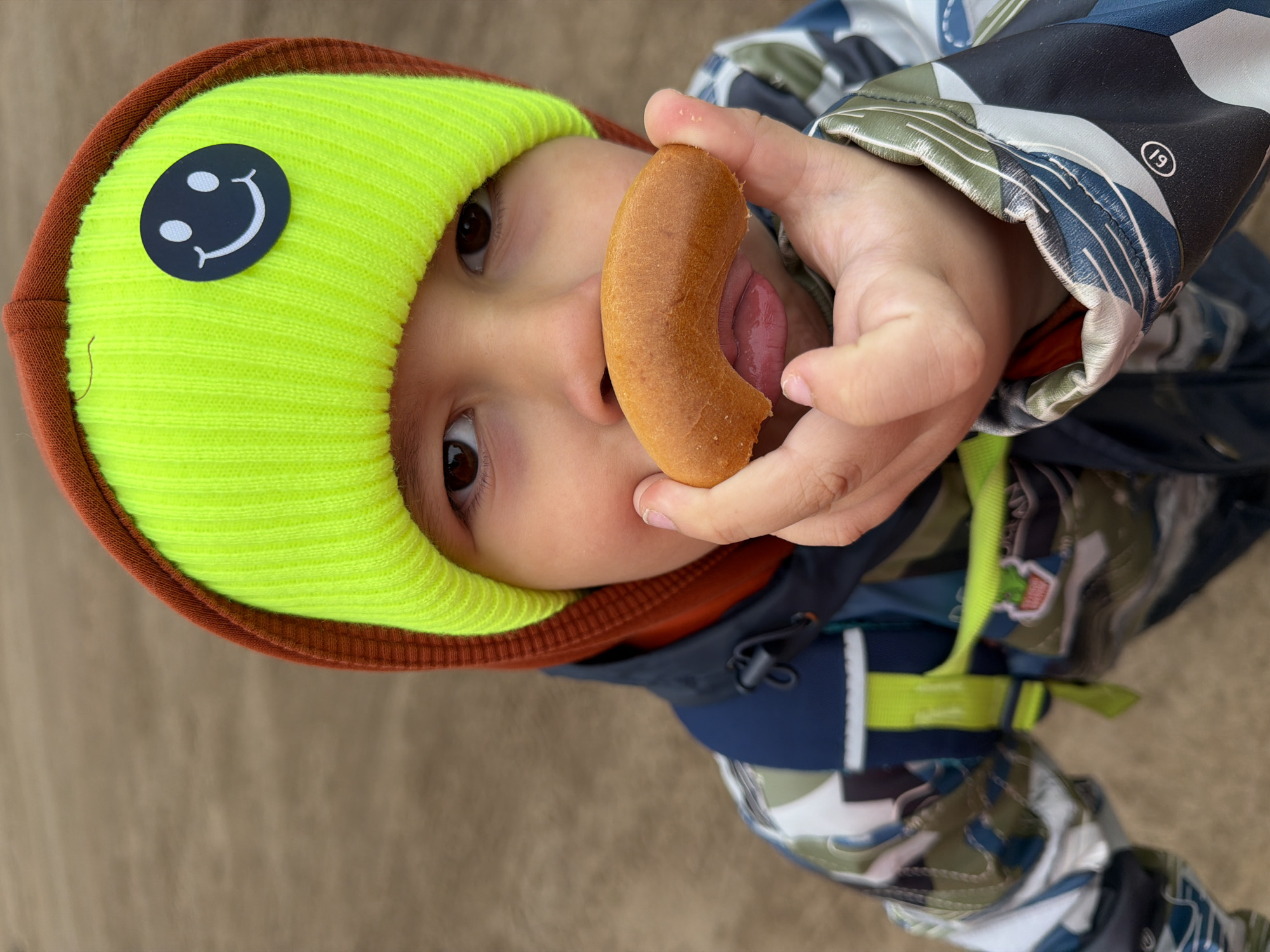 A toddler, wearing a bright yellow and brown hat with a smiley face patch, is holding a partially eaten cylindrical pastry up to their face and sticking their tongue out. The child is wearing a patterned jacket, possibly camouflage or geometric designs in blues, greens, and whites. The background appears to be a neutral-toned surface, possibly concrete or a wooden floor. Based on the information, the picture was taken in the city of Chaikovsky, Russia.