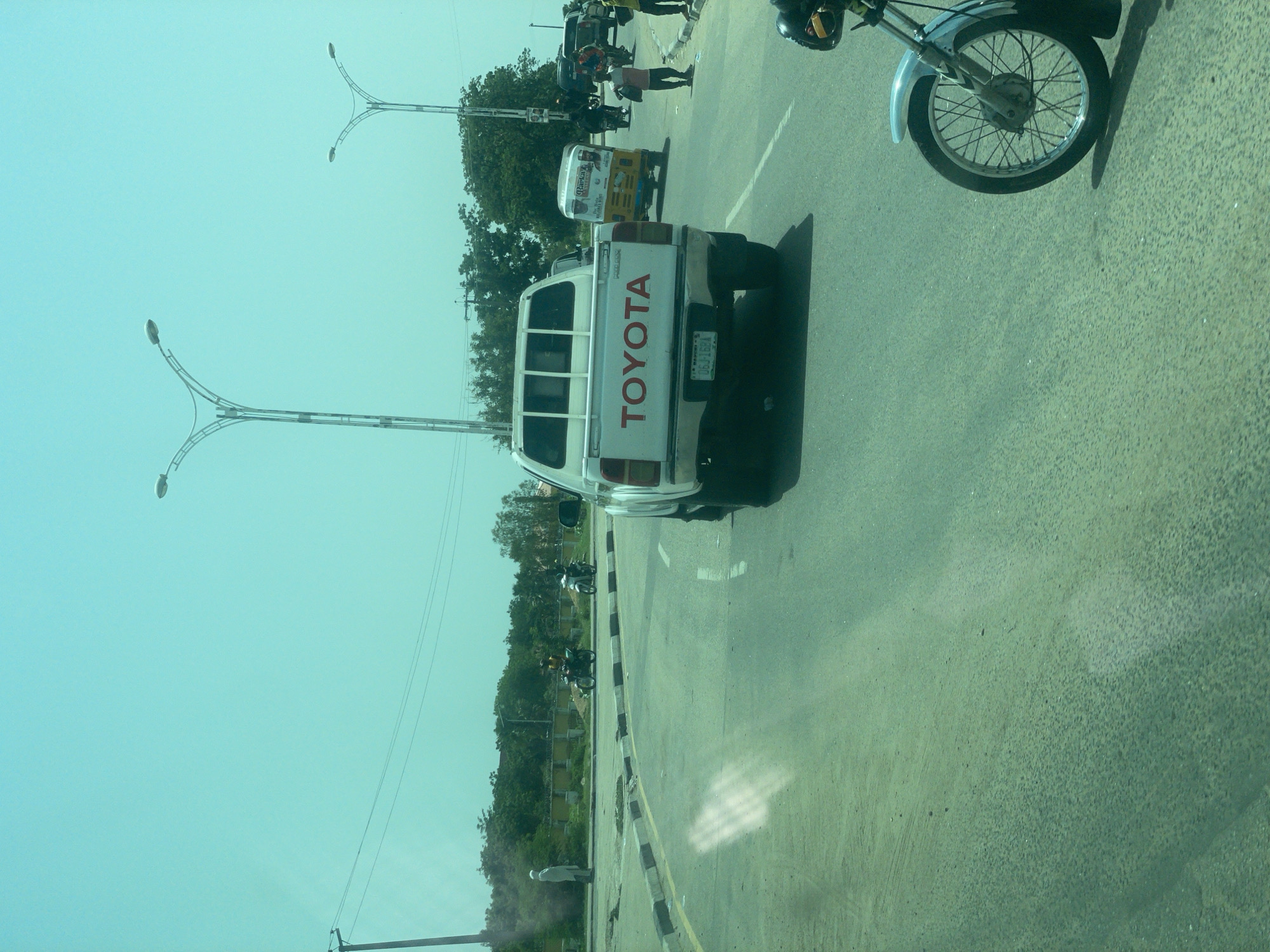 This media file captures a street scene in Bauchi, Nigeria, likely during the daytime given the bright lighting and clear sky, suggesting fair weather. The primary subject is a white Toyota pickup truck, prominently displaying "TOYOTA" in large red lettering on its tailgate. The truck is positioned on an asphalt road, with a curb and a grassy median visible to its left.
Several other vehicles are present, including a yellow auto-rickshaw with advertising on its side, and several motorcycles. One motorcycle, a lighter colored one, is partially visible in the foreground to the right, showing its wheel and fender. Another motorcycle is seen further down the road with two people on it. A person in a white shirt stands near the curb on the left side of the road. In the background, there are trees and vegetation, indicating a setting that is not purely urban.
Streetlights with multiple lamps on curved arms are visible, suggesting this is a street or intersection that is illuminated at night. Some overhead wires are also present. The overall atmosphere appears to be that of a busy roadside, possibly an intersection or a marketplace area given the variety of vehicles and the presence of advertising. No specific event or activity is clearly defined, but the scene depicts typical daily traffic and street life. The license plate on the Toyota truck is partially visible, reading "06JIGM".