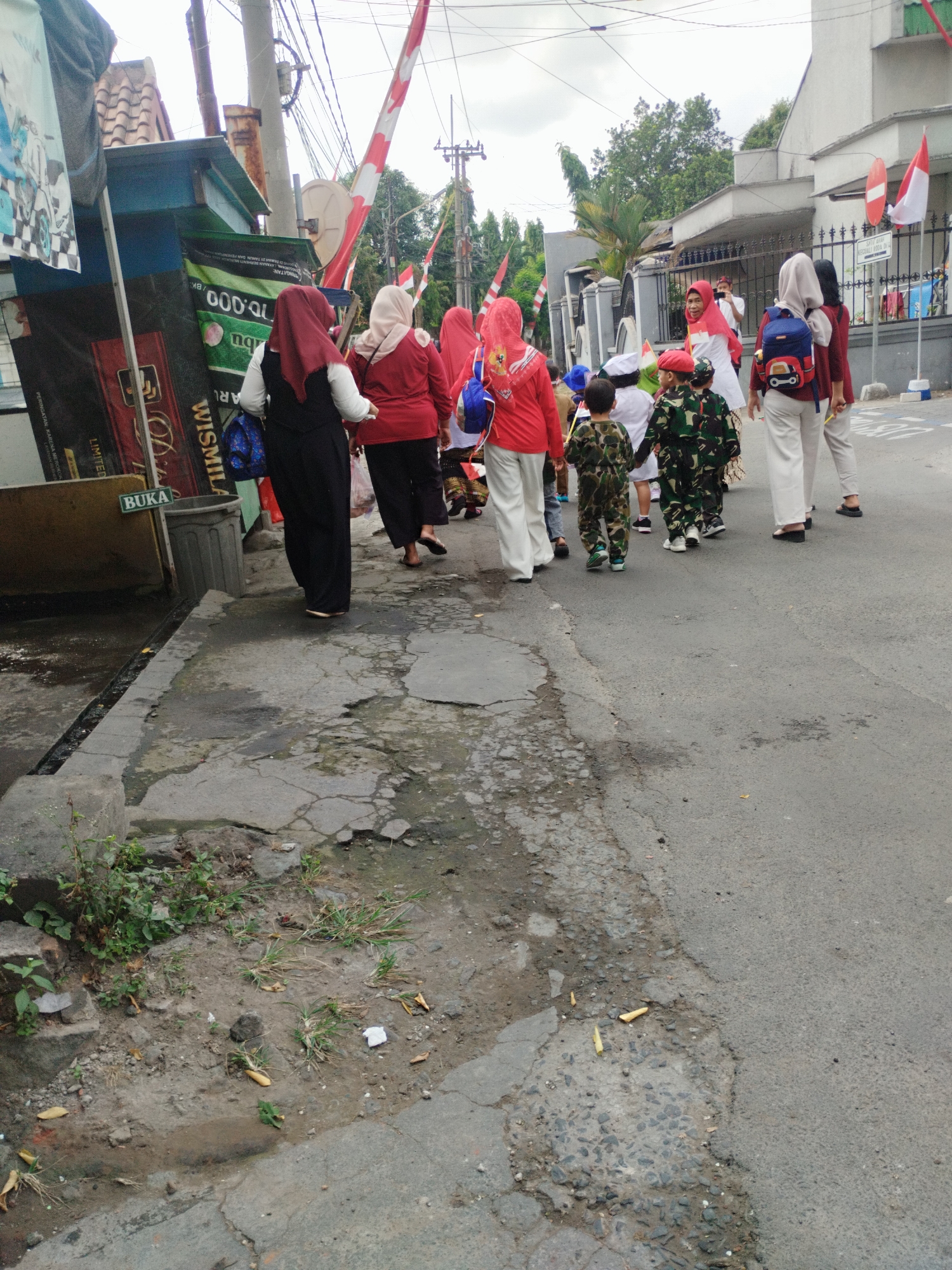 In Surabaya, Indonesia, a group of people, including children and adults wearing headscarves, walk along a paved street. The adults are dressed in red and white, while some of the children wear camouflage-patterned outfits. In the background, a white building with flags and a red and white striped pole are visible. The street is lined with buildings and a fence, and there are trees and power lines overhead. The pavement on the left side of the street is cracked and uneven, with some patches of dirt and vegetation. A trash can is also visible near the buildings. The scene appears to be a daytime outdoor gathering or parade.