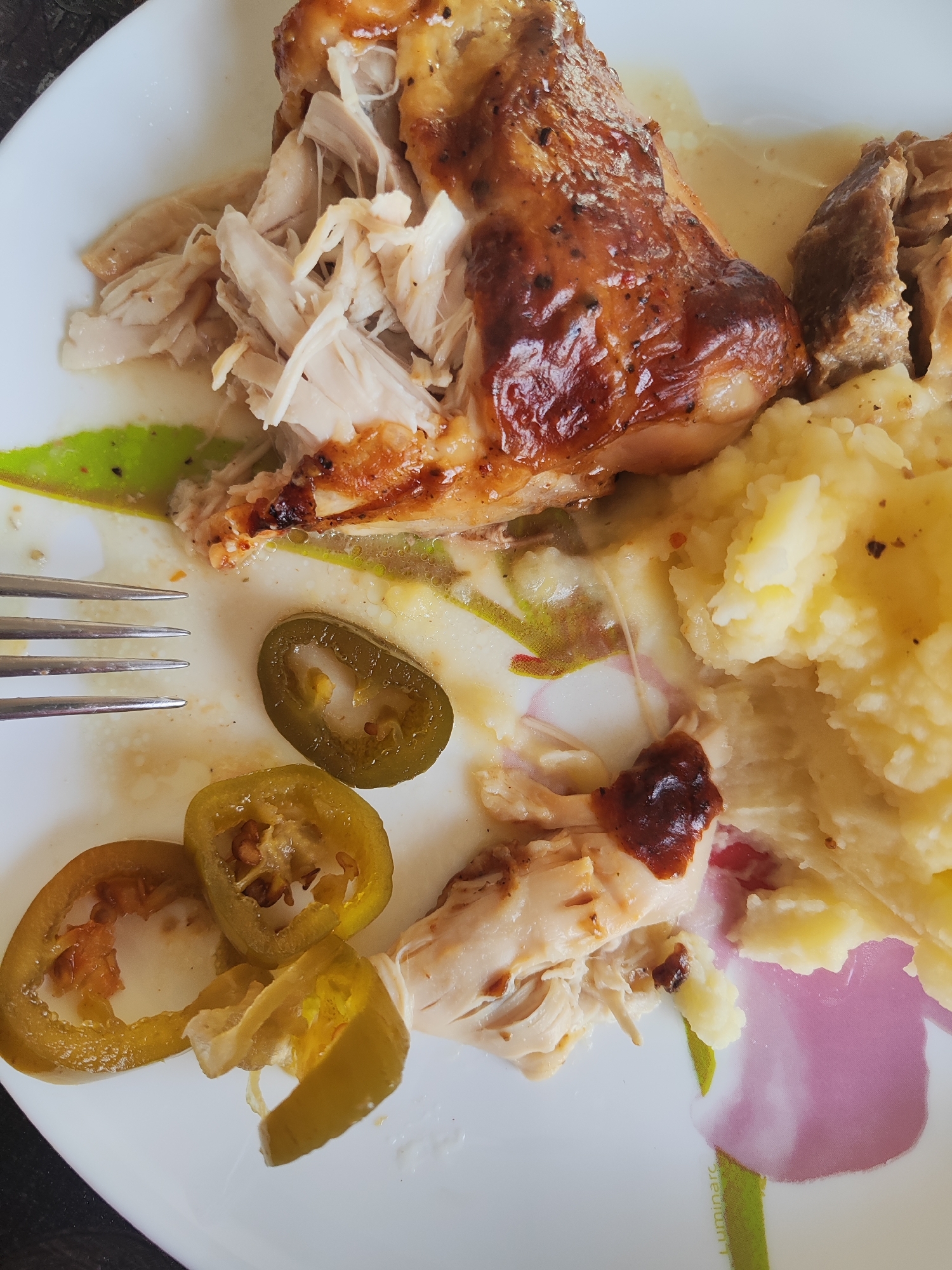 A close-up, overhead shot shows a white plate with a floral design, holding a meal. The plate contains shredded cooked chicken, a piece of roasted chicken leg with crispy skin, a serving of mashed potatoes, and several slices of pickled jalapeños. A fork rests on the left side of the plate, with its tines partially obscuring some of the food. The meal appears to be partially eaten, with sauce and food remnants scattered across the plate. The background is dark and out of focus. There is no discernible information about the location in Gvardeysk, Russia, or any people or actions in the image.