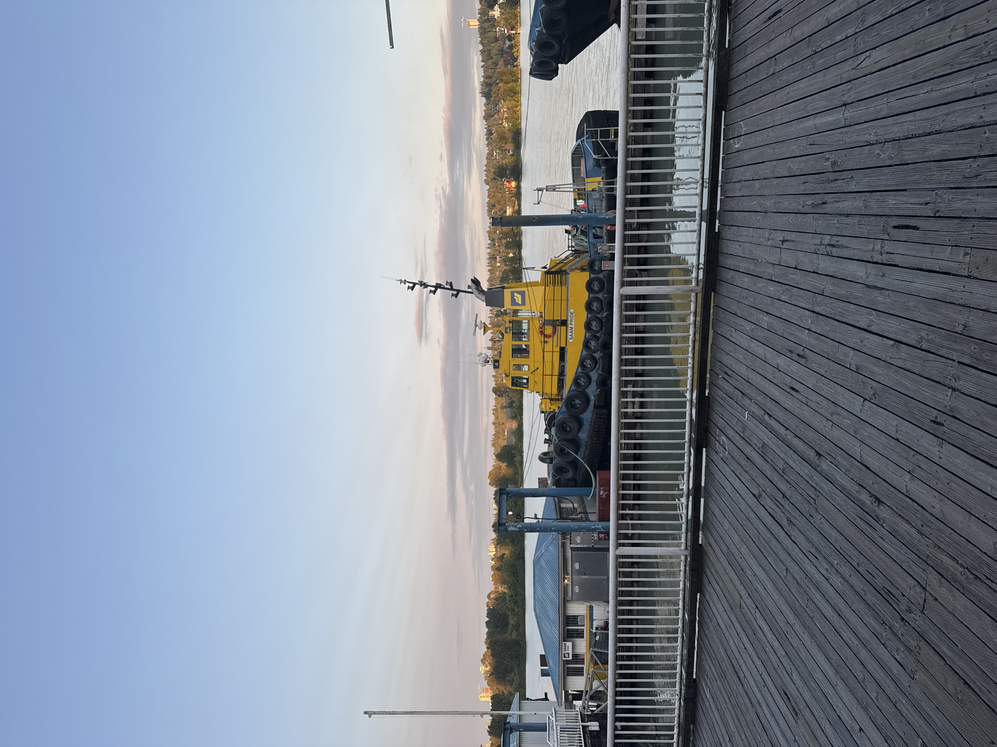 A wide shot captures a yellow tugboat docked at a pier on the Fraser River in New Westminster, Canada. The tugboat, named "SAAM PRIDE," is equipped with a black bumper and is tied to a small building with a blue roof. The pier is made of weathered wooden planks and has a white metal railing. In the background, trees and buildings line the opposite bank of the river under a pale blue sky with wispy clouds. The water is calm and reflects the sky and surrounding elements. The scene is serene and appears to be a peaceful moment at the waterfront.