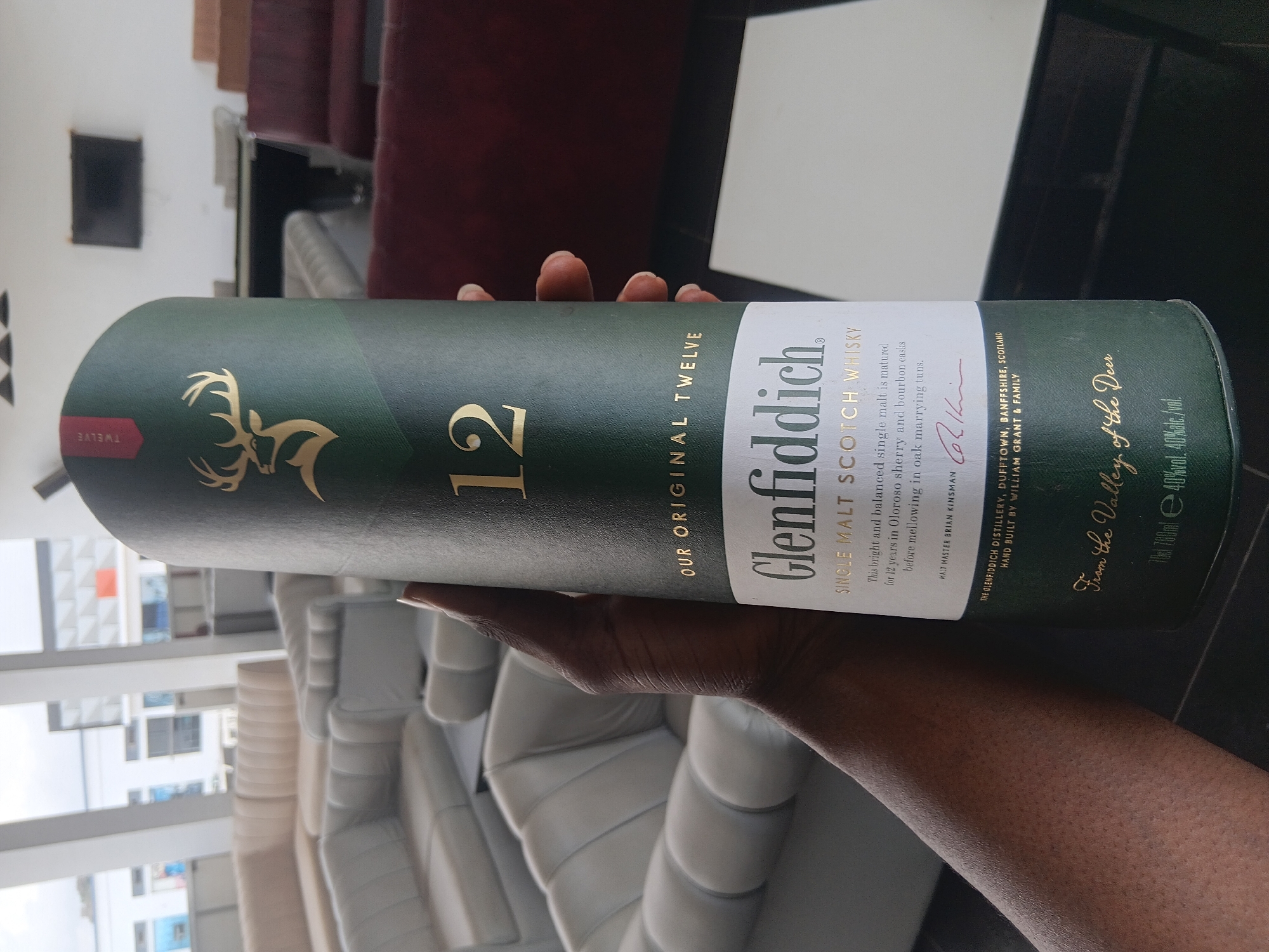 A person is holding a container of Glenfiddich 12 single malt Scotch whisky. The container is dark green with gold lettering and a gold stag emblem. The person's hand and part of their arm are visible. The background includes a blurry interior scene with furniture, possibly a sofa, and a window with an exterior view. The location appears to be indoors but the exact location in the city of Gura Top, Nigeria is impossible to determine without additional information.