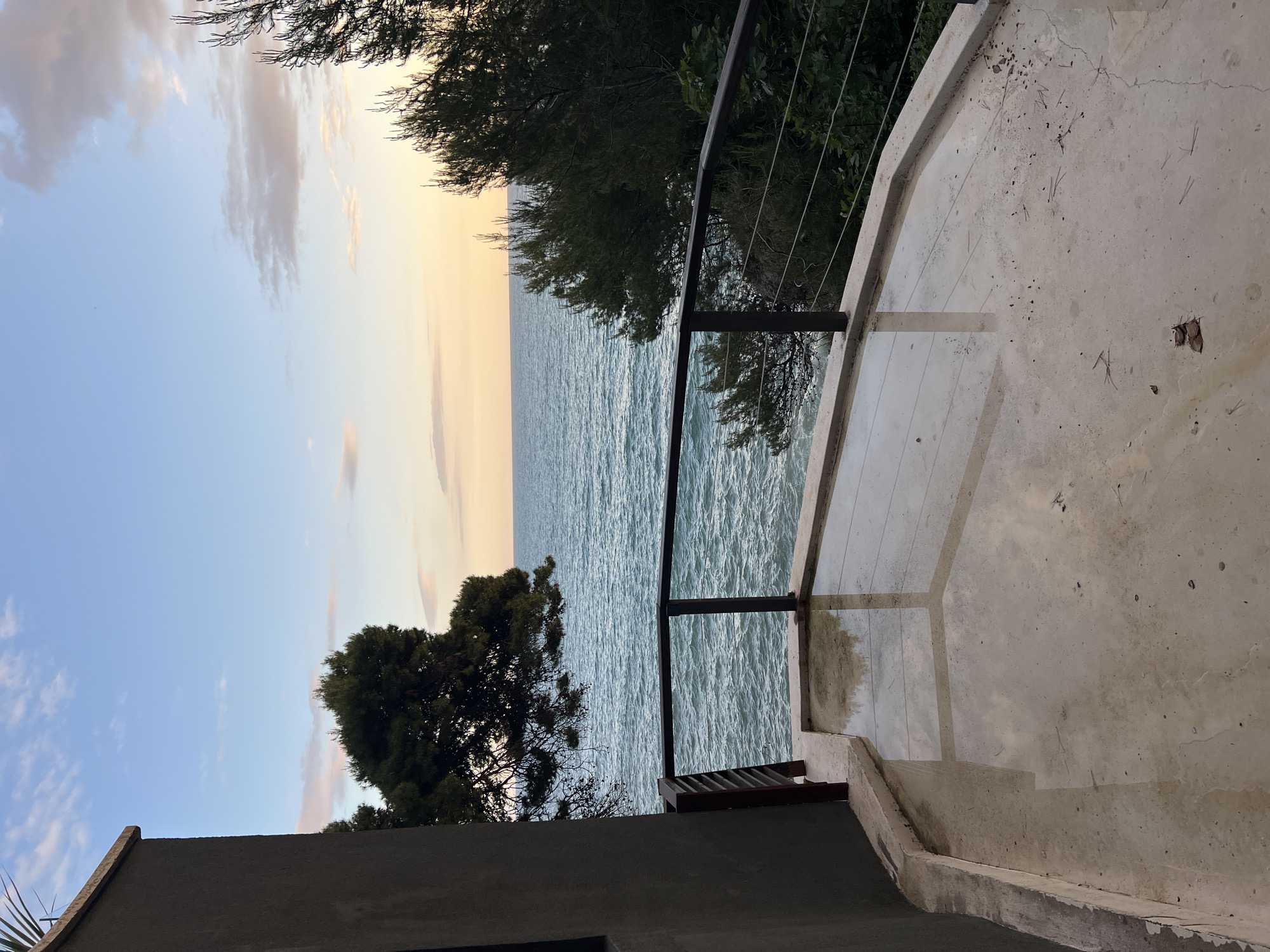 The image captures a coastal scene in Ulcinj - Ulqin, Montenegro, as seen from an elevated, concrete patio. A metal railing borders the patio's edge, partially obscured by foliage from nearby trees. Beyond the railing lies the sea, its surface ruffled by gentle waves, reflecting the diffused light of the sky. The sky transitions from blue to a light orange/yellow hue, indicating either sunrise or sunset. Trees are positioned along the shoreline. A portion of a building, possibly the structure the patio belongs to, is visible in the lower part of the image.