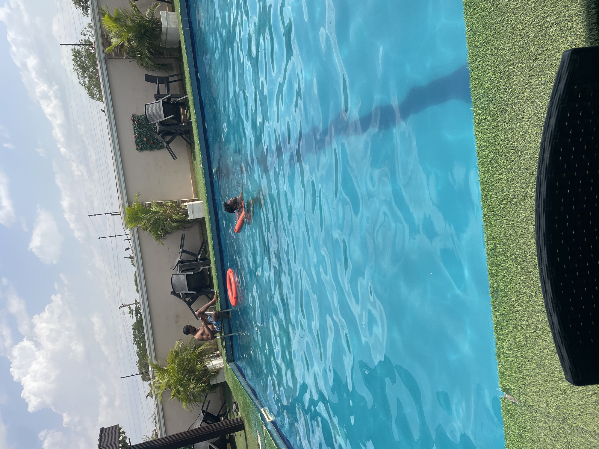 This image captures an outdoor swimming pool scene in Abuja, Nigeria, under bright daylight. The clear blue water of the pool is the central element, reflecting the sky and showing ripples on its surface.
Two individuals are visible enjoying the pool. One person is partially submerged, floating or swimming with an orange inflatable ring around their torso. Closer to the foreground, another person is sitting on the steps or edge of the pool, with their legs in the water, wearing patterned swim shorts.
The poolside is lined with green artificial turf, separating the water from a light-colored building wall. Along this wall, several black outdoor chairs, some with a modern metal frame and others with a woven texture, are arranged, interspersed with small palm trees and other green foliage. In the background, beyond the building, additional trees and utility poles with power lines are visible against a bright, cloudy sky, indicating a clear and sunny day. A dark, possibly woven, outdoor chair or table is partially visible in the bottom right corner, suggesting the viewer's immediate surroundings.
The scene conveys a relaxed and leisurely atmosphere. There is no visible text in the image.