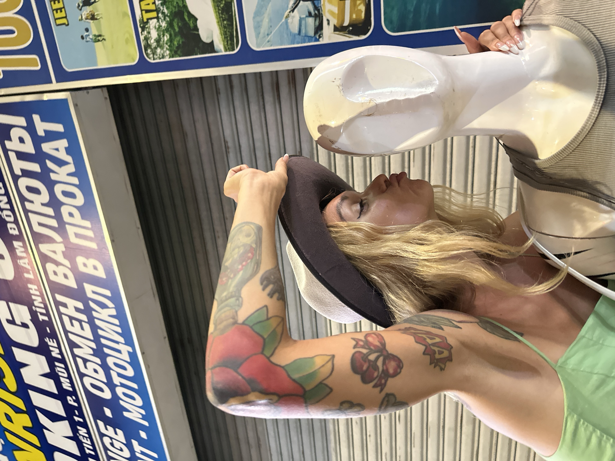 A young woman with blonde hair, wearing a light green tank top and a wide-brimmed hat, leans in to kiss a white mannequin in Phu Thuy Ward, Vietnam. Her right arm, covered in colorful tattoos, is raised, and her left hand is placed on the mannequin's neck. A blue and yellow sign above them reads "WRISTKING" and other text in Vietnamese and Russian, likely advertising currency exchange and rentals for motorcycles and ATVs. The background consists of a closed metal roll-up door.