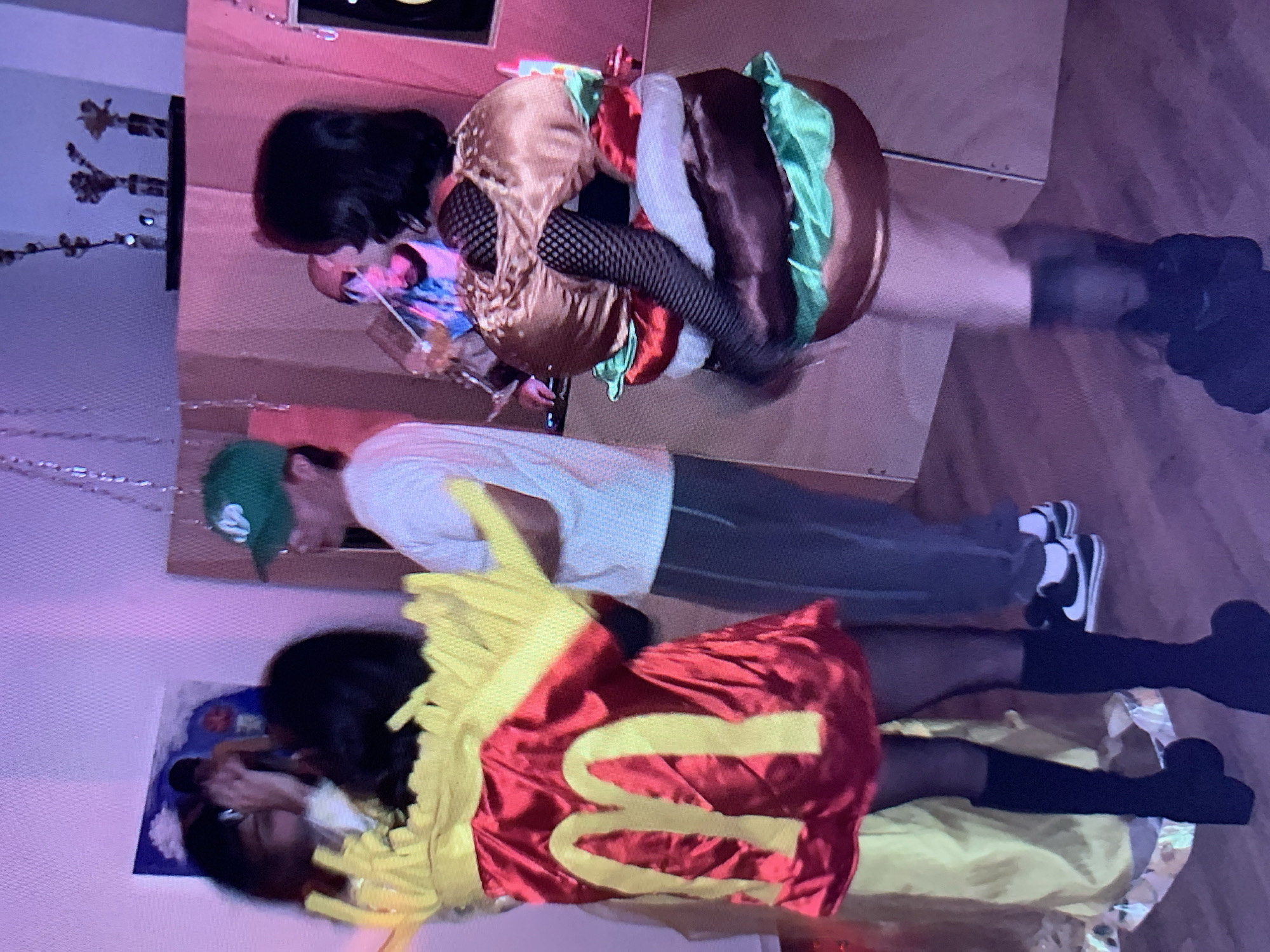 The image shows three people posing indoors. One person is dressed in a hamburger costume, another in a French fries costume, and a third is wearing a white t-shirt, grey pants, a green hat, and black and white sneakers. The environment appears to be a room with a casual setting and possible decorations. The image was likely taken indoors. There is no way to know if this image came from Tel Aviv, Israel.