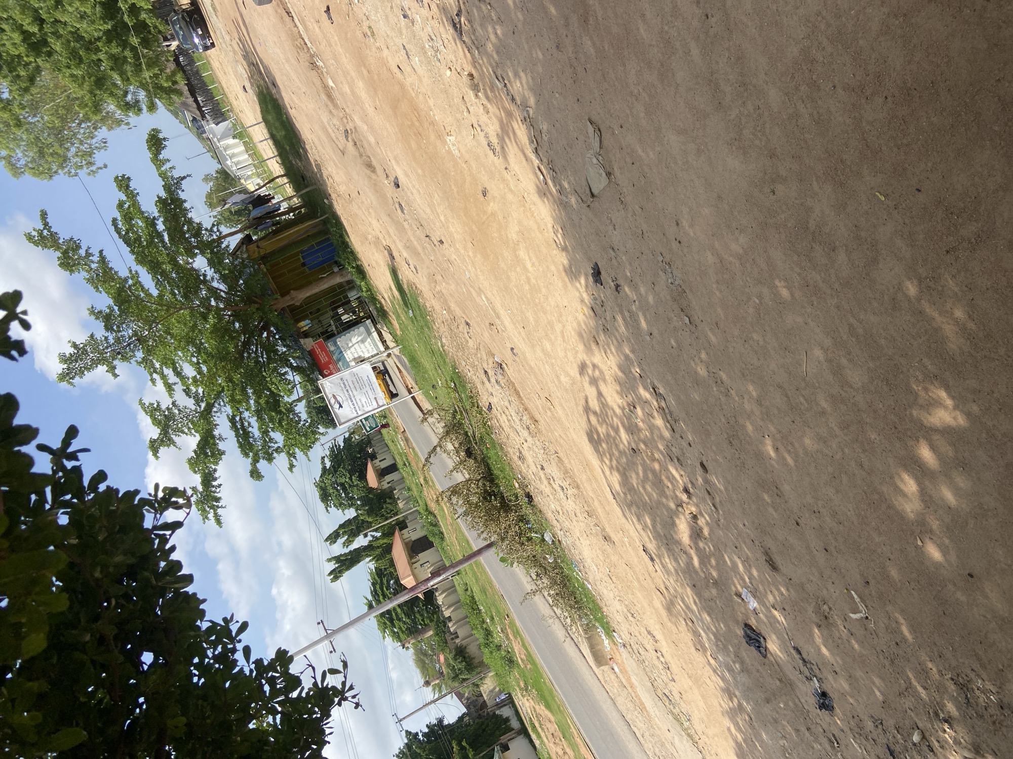 This image depicts a roadside scene in Jalingo, Nigeria, on a sunny day with scattered clouds. The foreground is dominated by a dirt and gravel ground with sparse vegetation and scattered debris, including plastic bags and papers. In the mid-ground, a paved road runs diagonally from the lower left to the upper right of the frame. Along the roadside, there are several structures, including a small, brightly colored building with clothes hanging to dry outside, and a signpost with text in English. Further back, a wall encloses properties with houses visible behind it. Mature trees with green foliage are present throughout the scene, some casting dappled shadows on the ground. A dark-colored car is parked on the right side of the road. A single utility pole with wires is also visible. The overall impression is one of a somewhat undeveloped urban or peri-urban environment.
